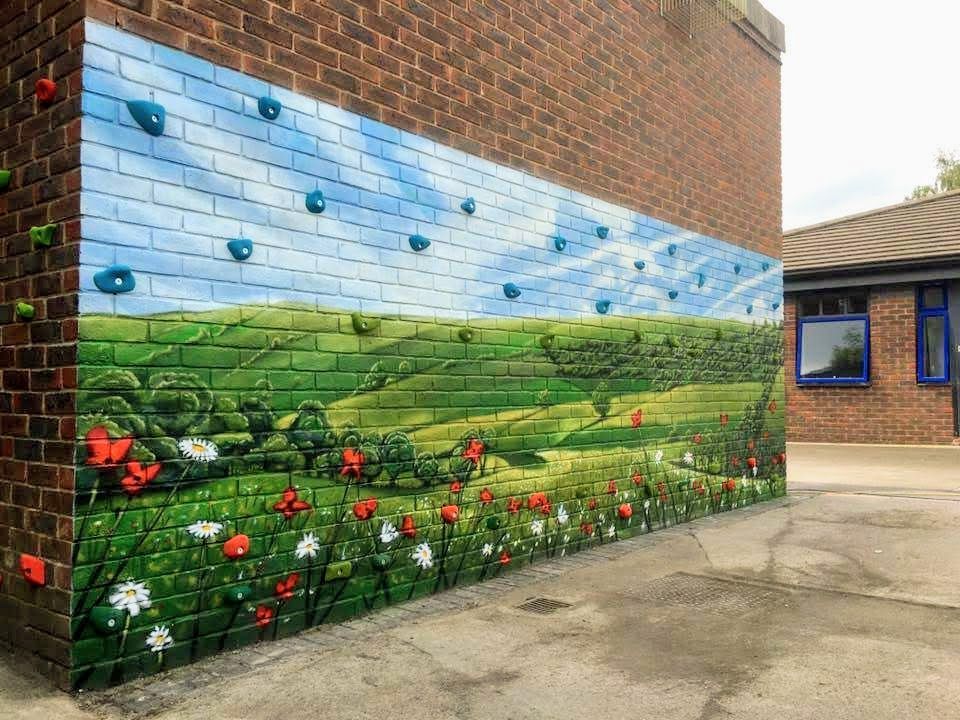 Mural of a landscape painted on a brick wall with climbing holds. Blue sky, green fields, red and white flowers.
