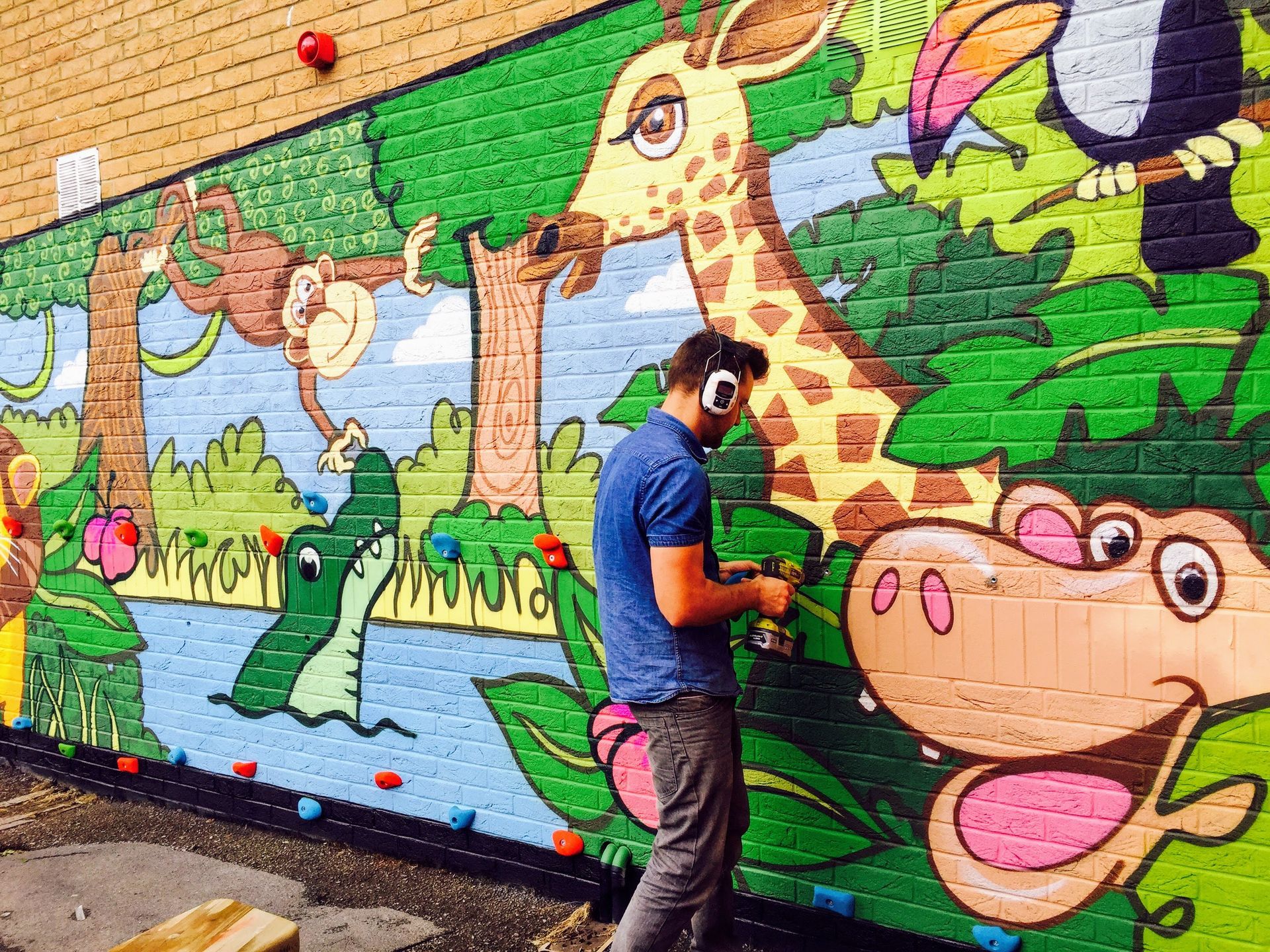 Man painting a jungle mural of animals on a brick wall, including a giraffe, monkey, and crocodile.