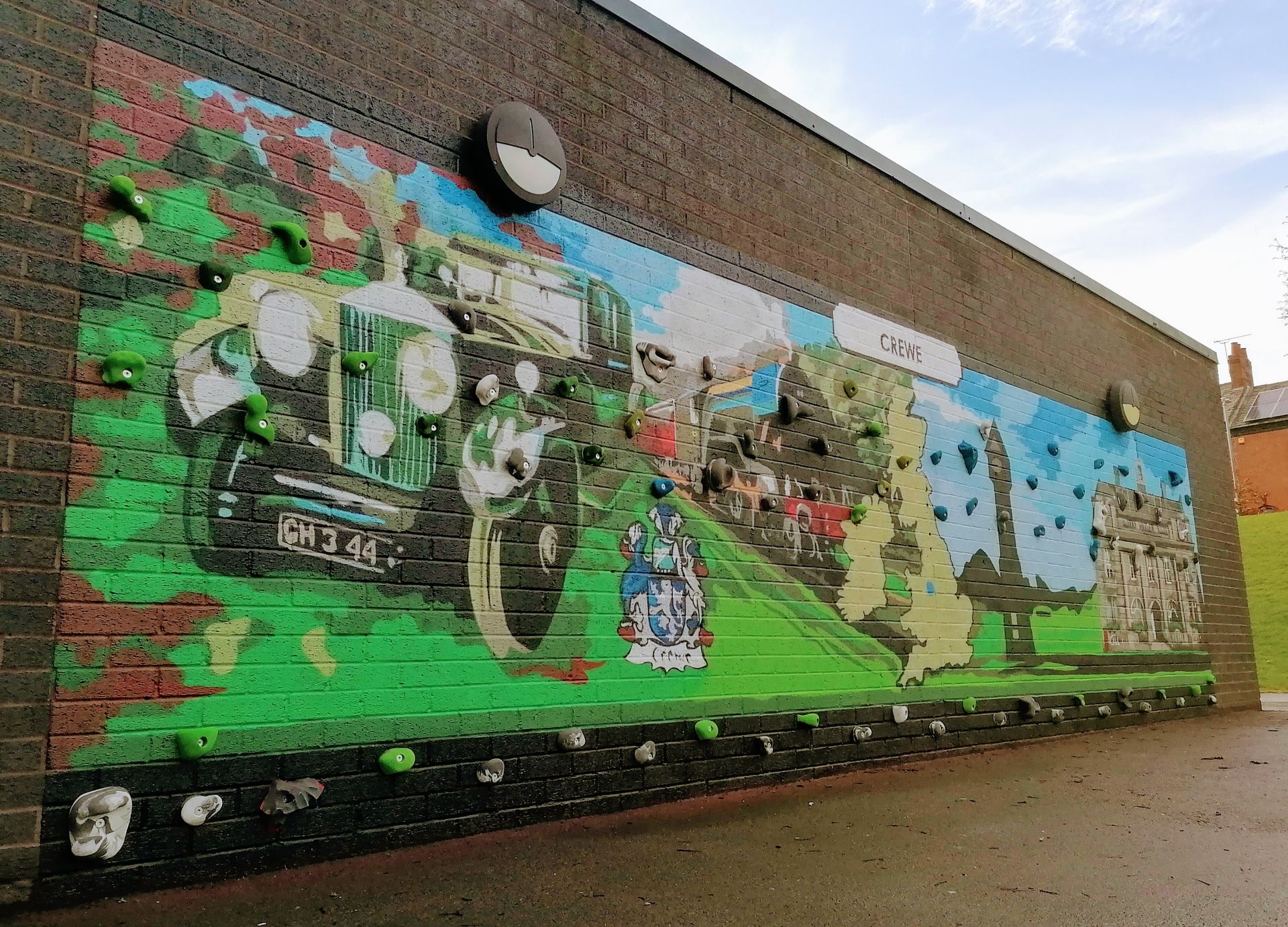 Mural on brick wall, depicting a vintage car, figures, and scenery. Climbing holds are embedded.