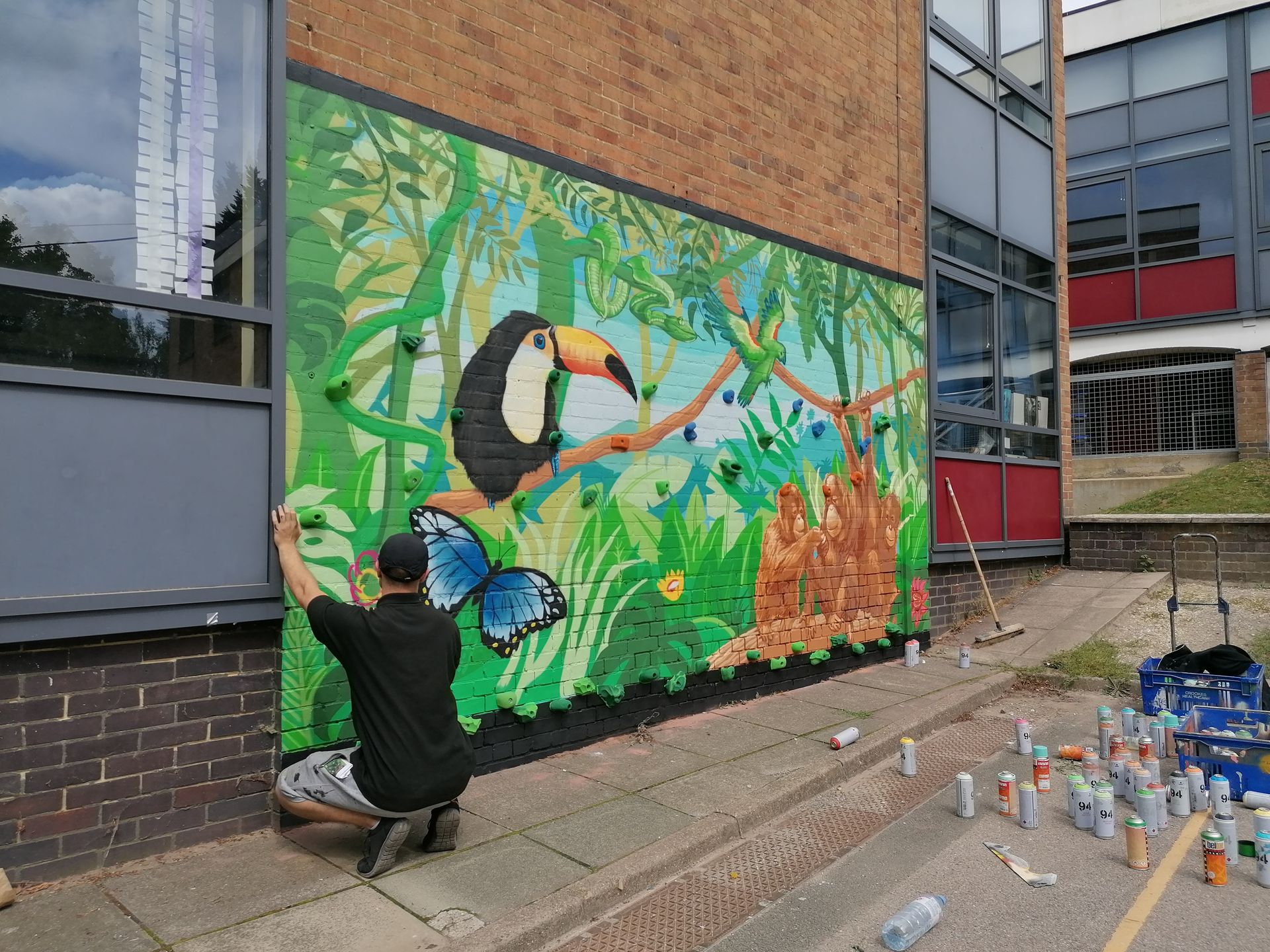 Man painting a jungle mural with a toucan, butterfly, and monkeys on a brick building exterior.