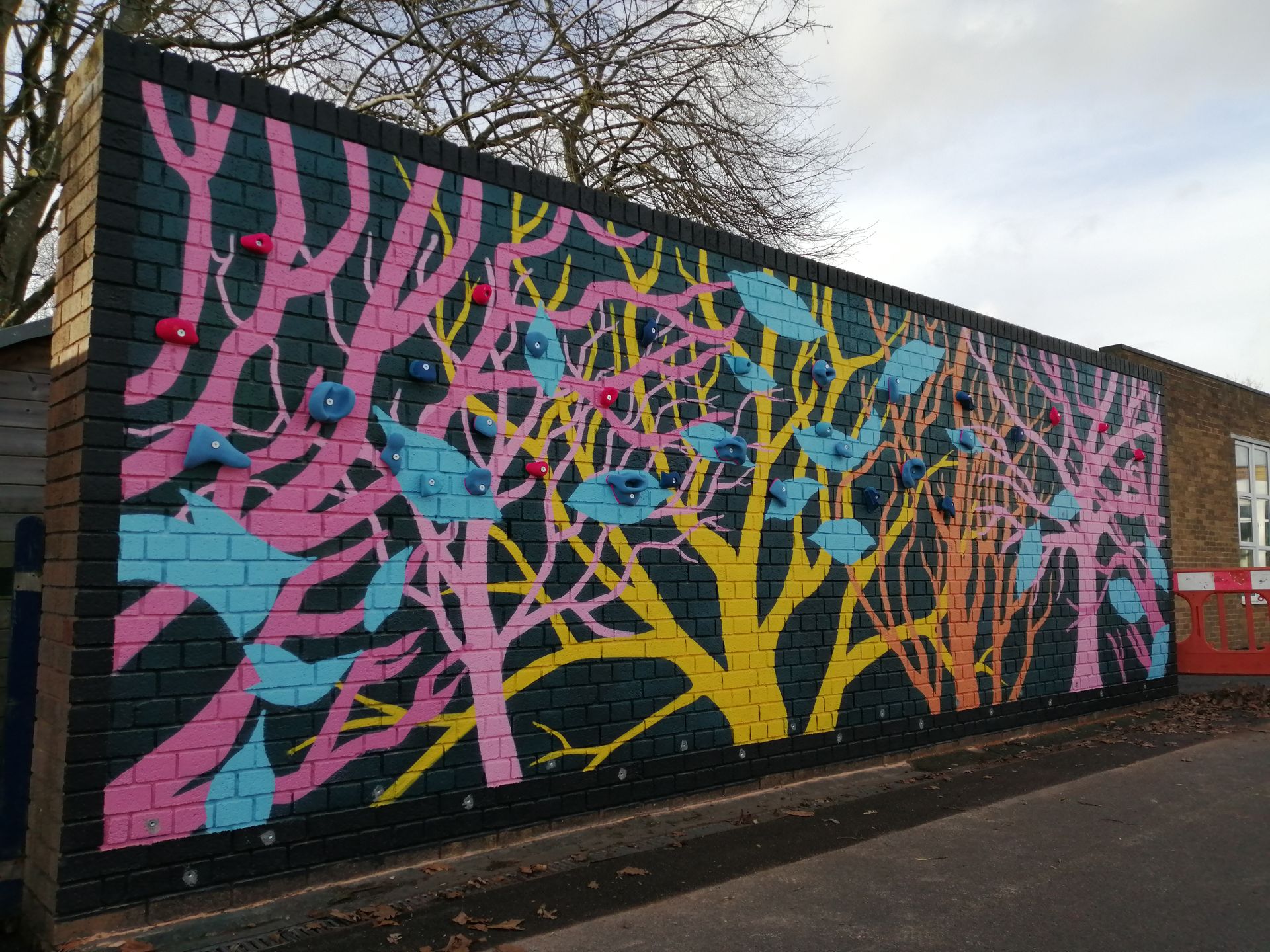 Colorful mural on a brick wall, depicting trees and birds. Pink, blue, yellow paint. Climbing holds attached.