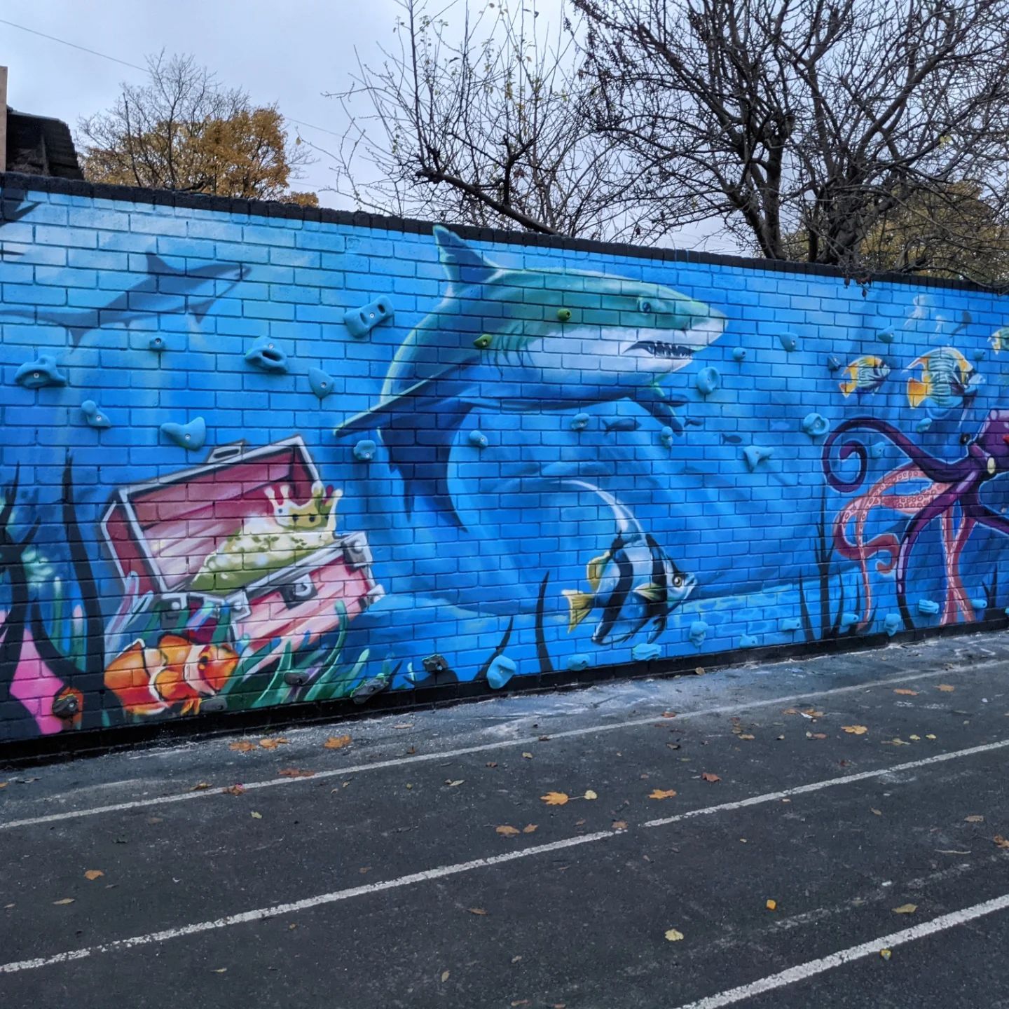 Mural of an underwater scene with a shark, fish, treasure chest, and octopus on a brick wall.