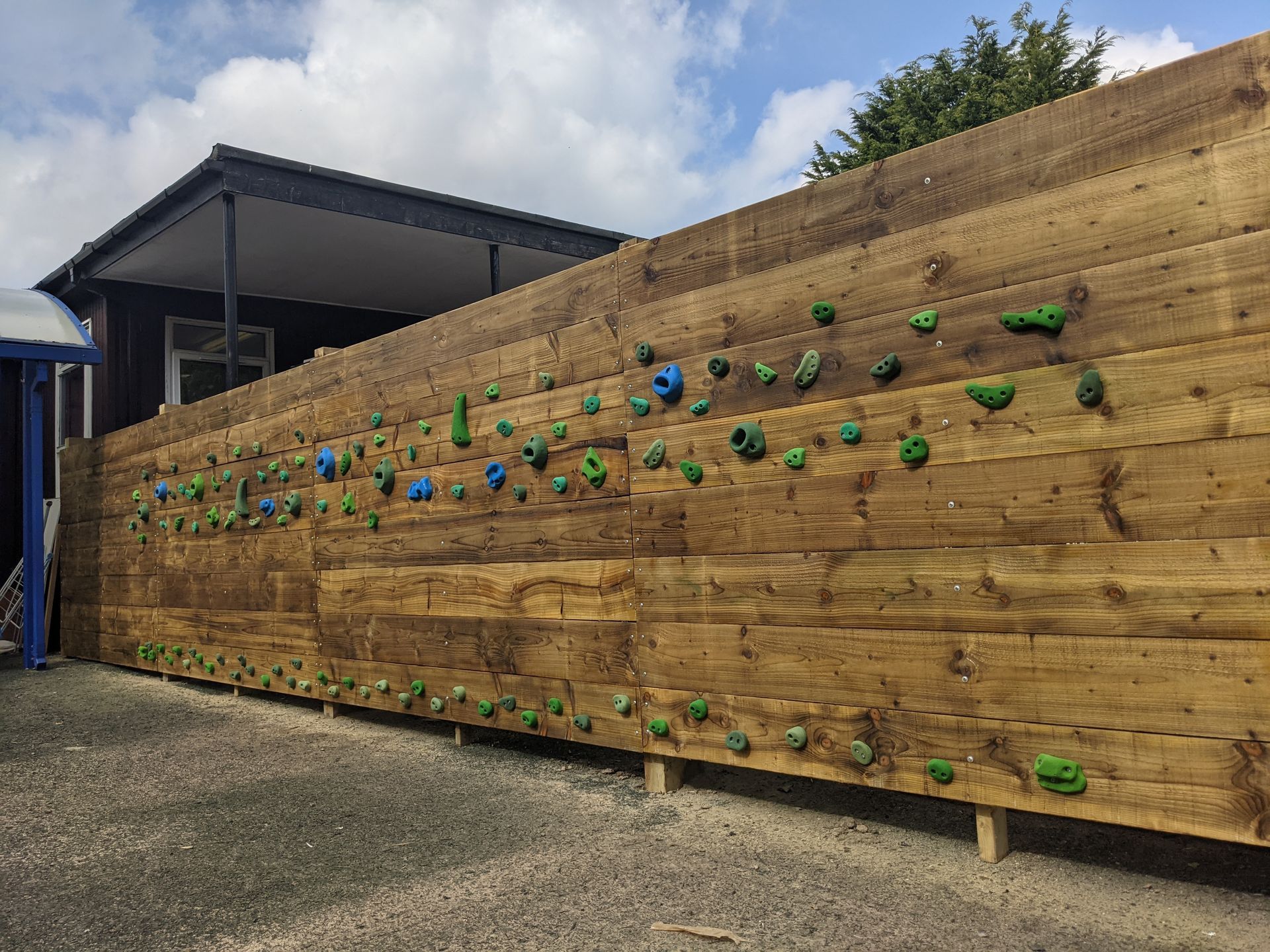 Wooden climbing wall with colorful handholds, outside a building, against a cloudy sky.