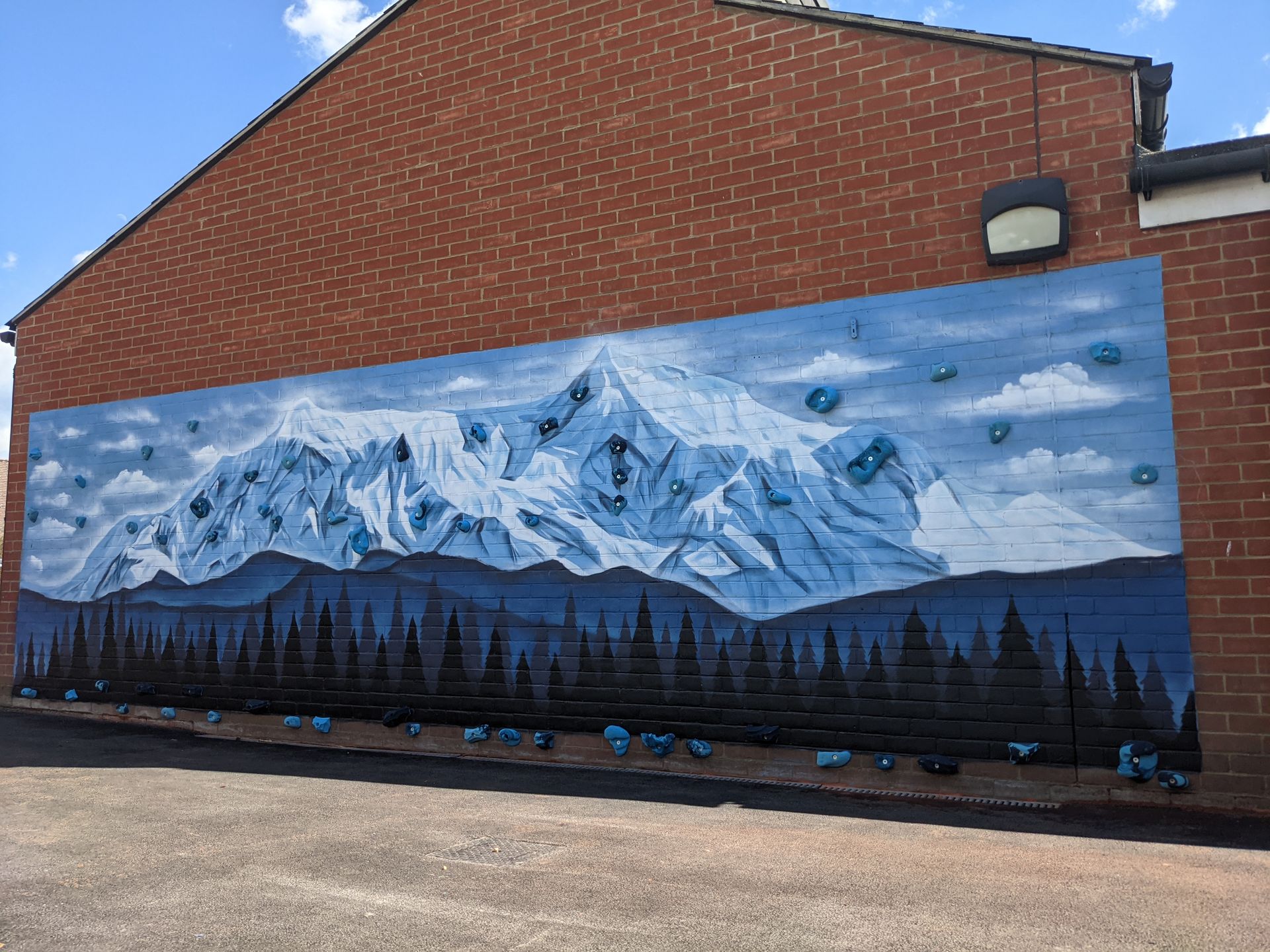 Mural of snow-capped mountains over a dark forest, painted on a brick building under a blue sky.
