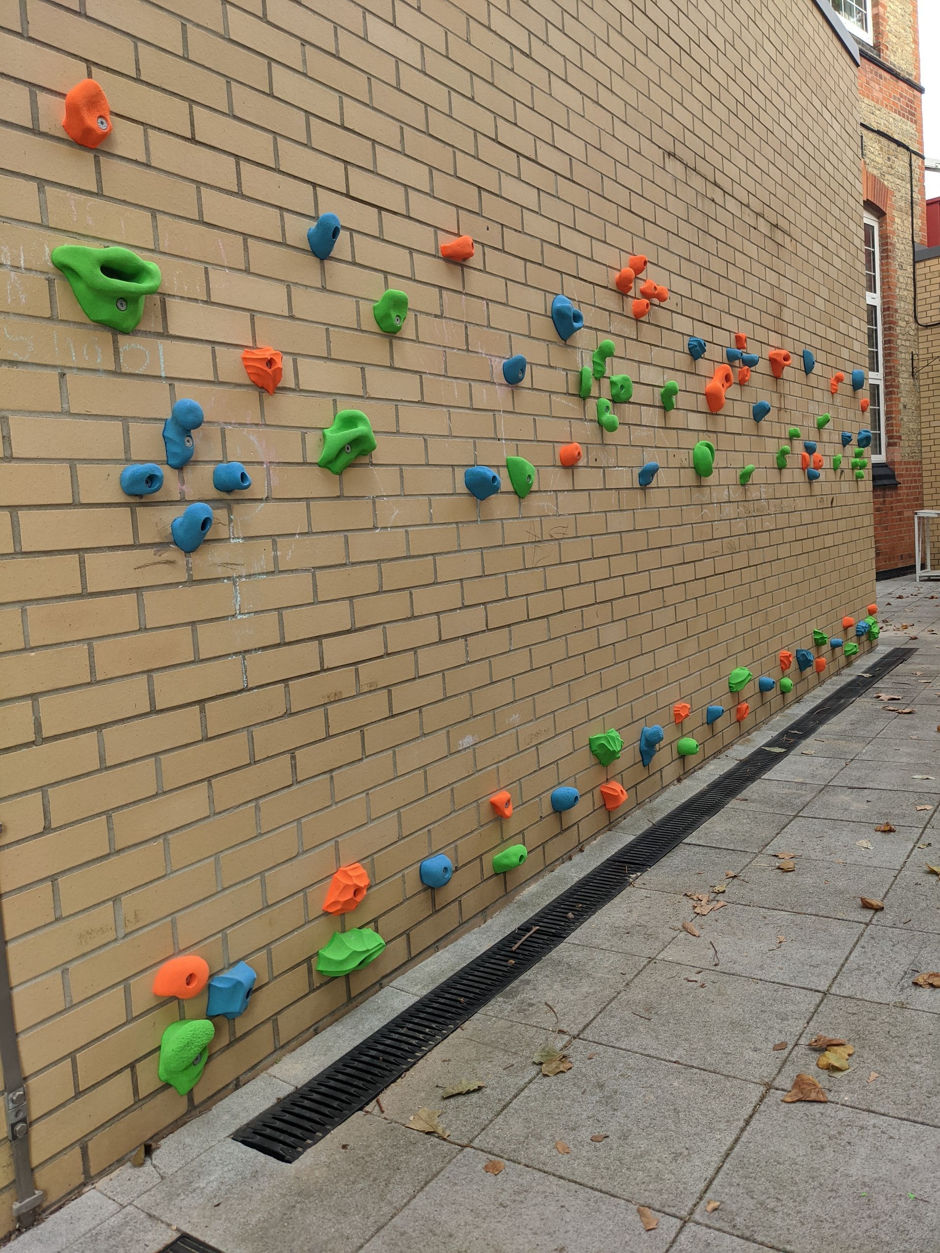Climbing wall with colorful holds on a brick building exterior, near a paved area.