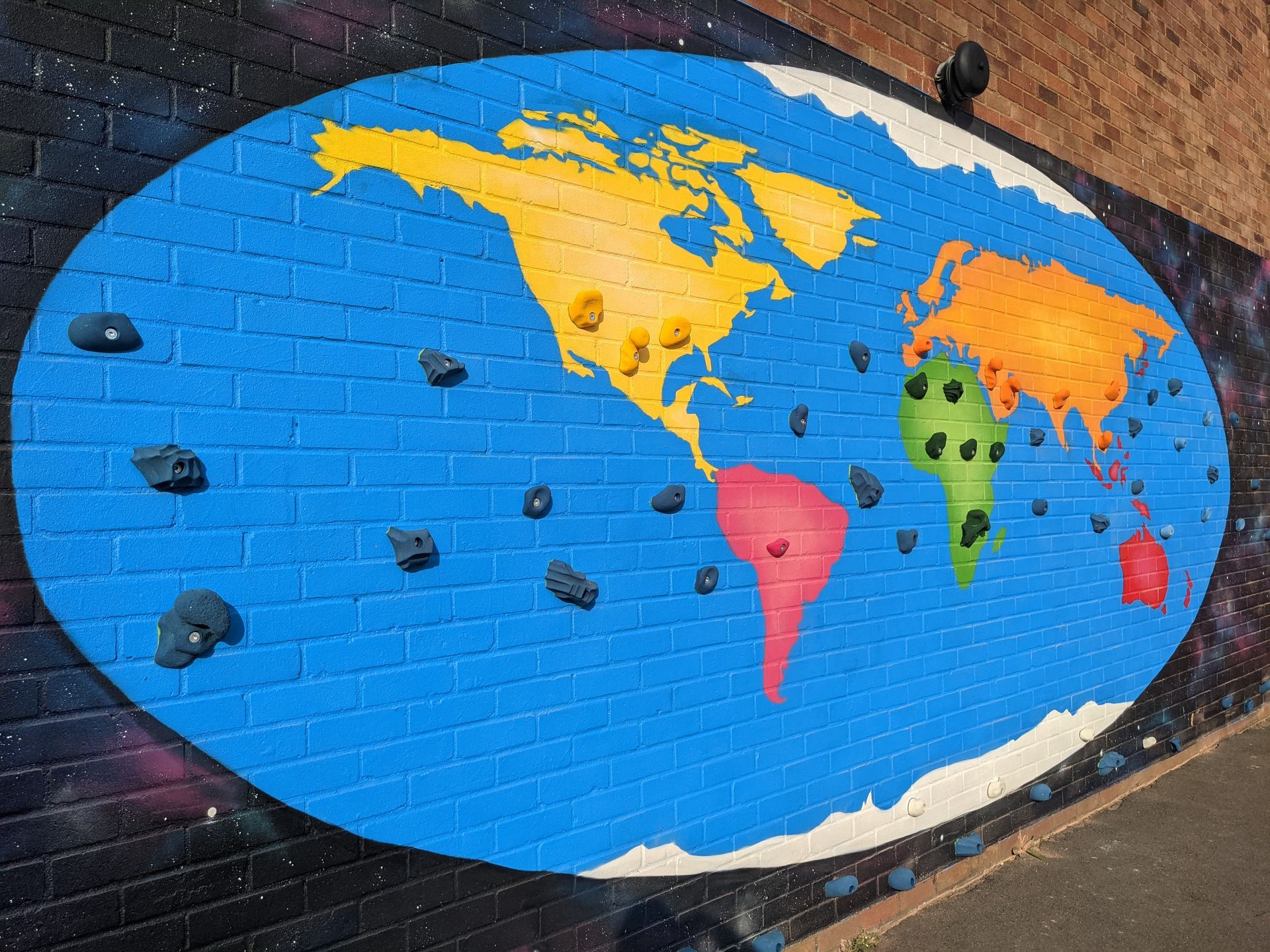 A colorful, oval map of the world painted on a blue brick wall, with climbing holds.
