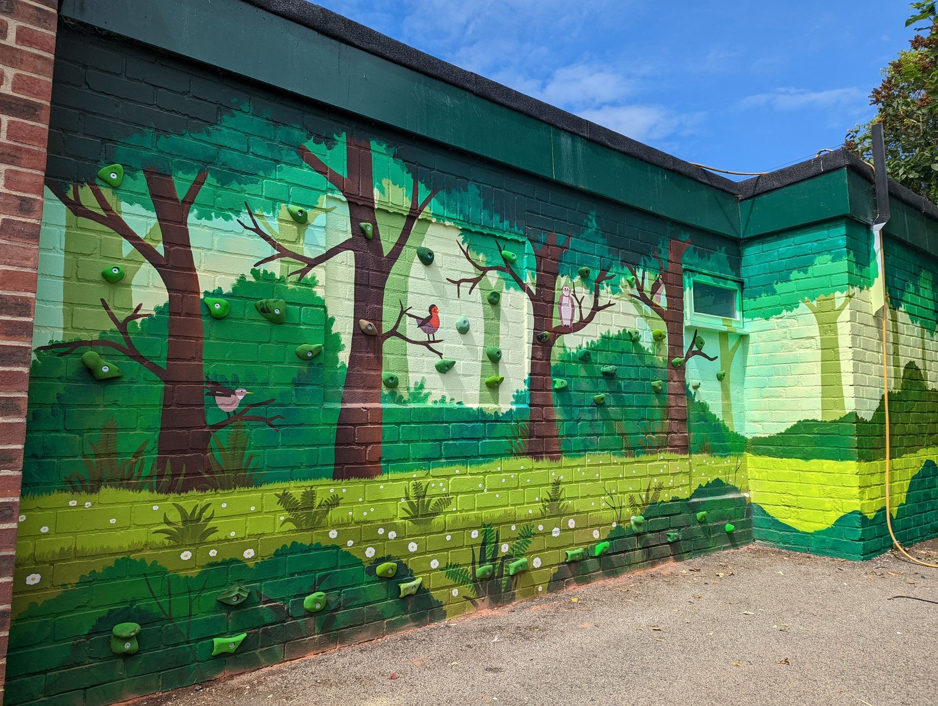 Mural of a forest scene on a brick wall with climbing holds. Green trees and foliage with birds and flowers.