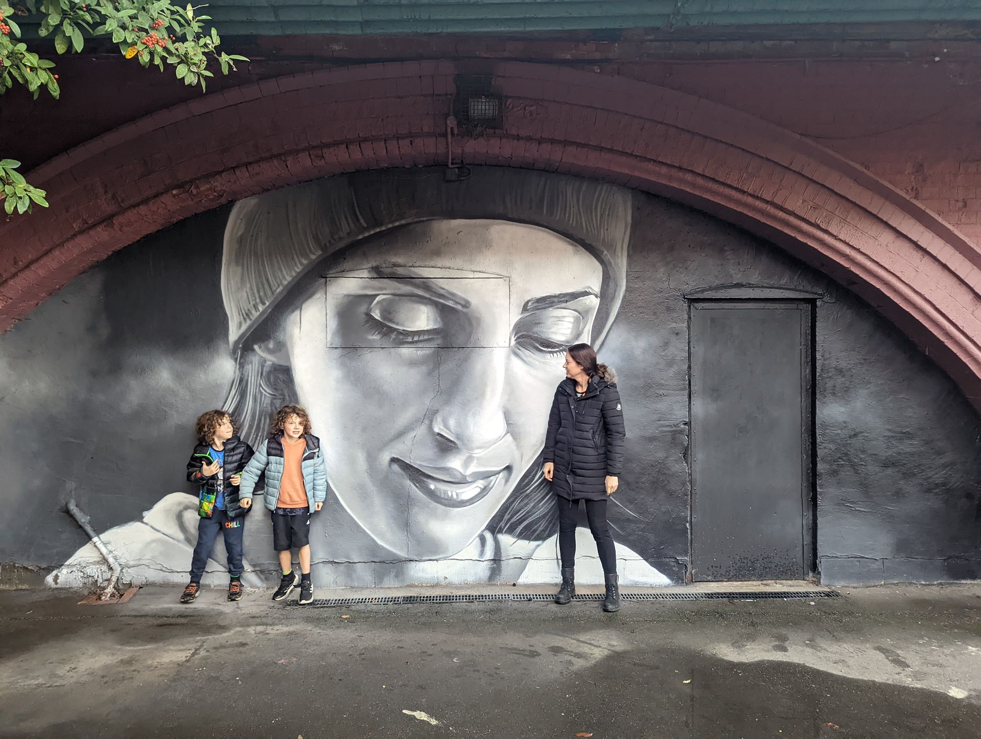Family poses in front of a large black and white mural of a person's face under a bridge.