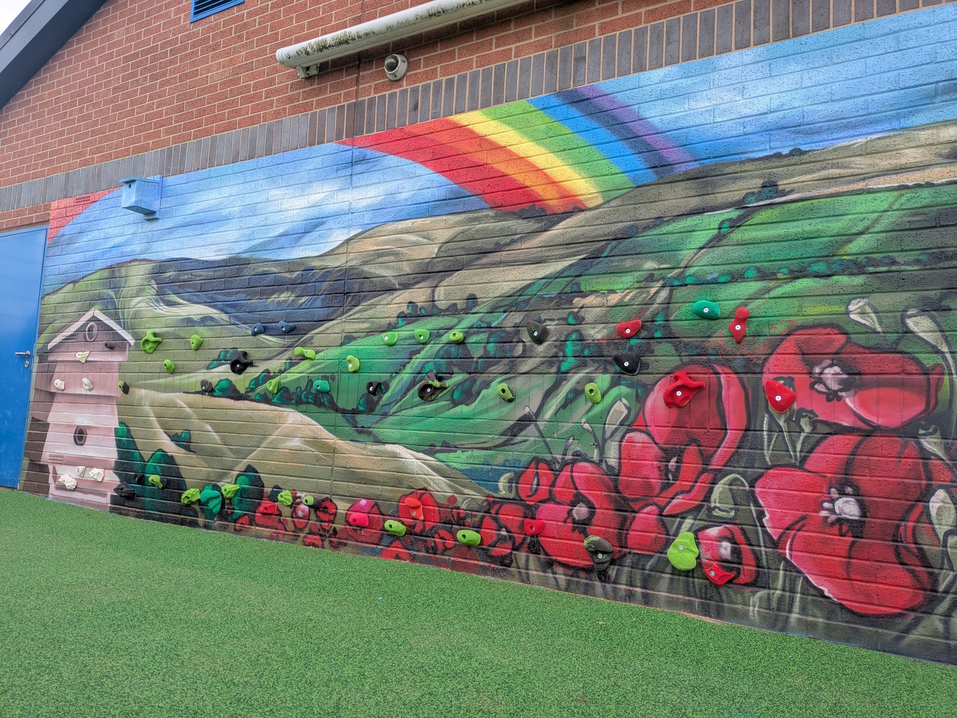 Mural on brick wall depicting a landscape with rainbow, mountains, flowers, and climbing holds.