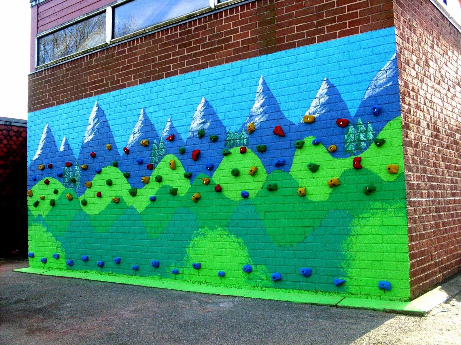 Painted climbing wall with blue and green mountain scene and colorful holds on a brick building.