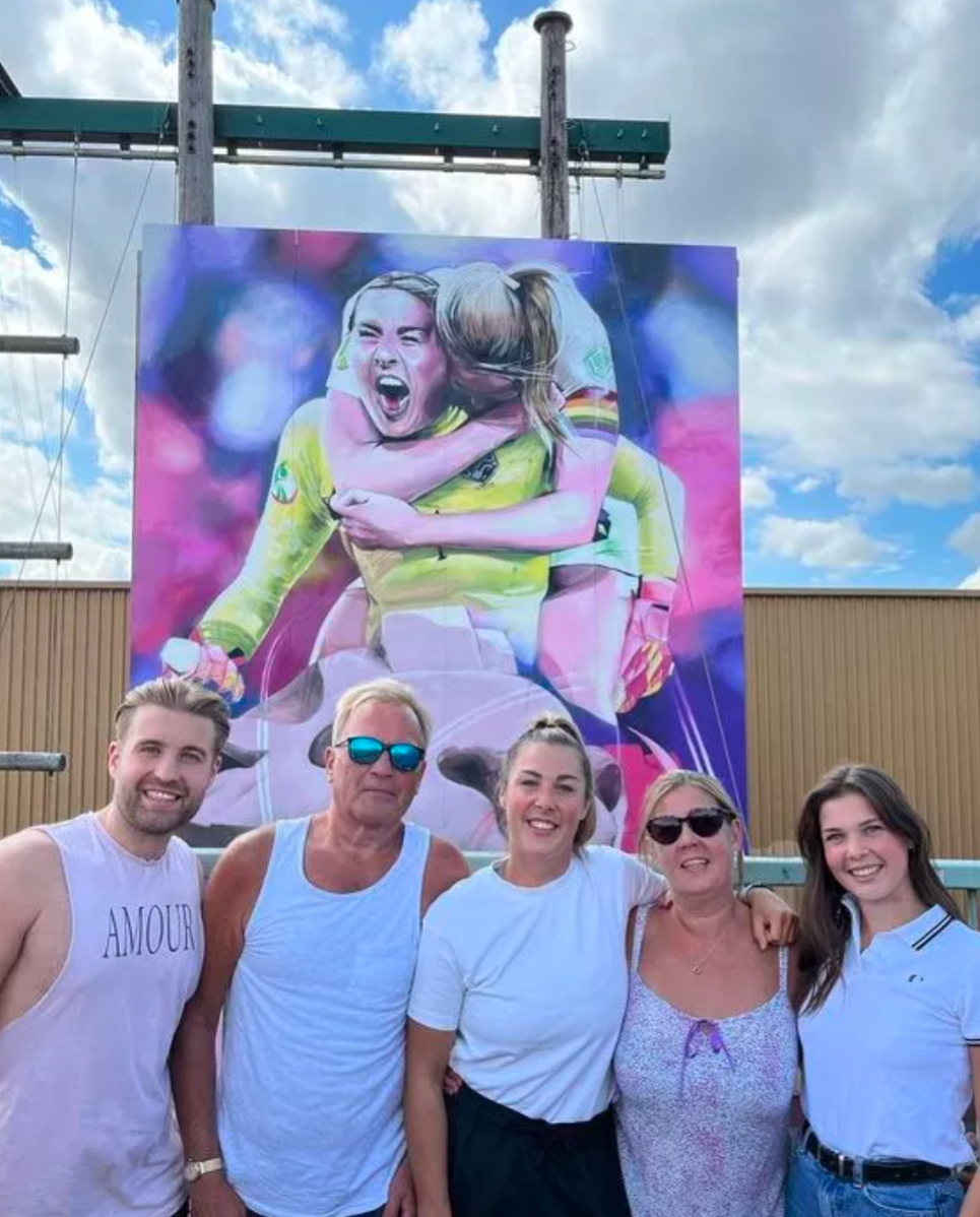 Two men pose in front of a sports-themed mural. One man holds his arm, the other smiles. The mural has two women and a lion.