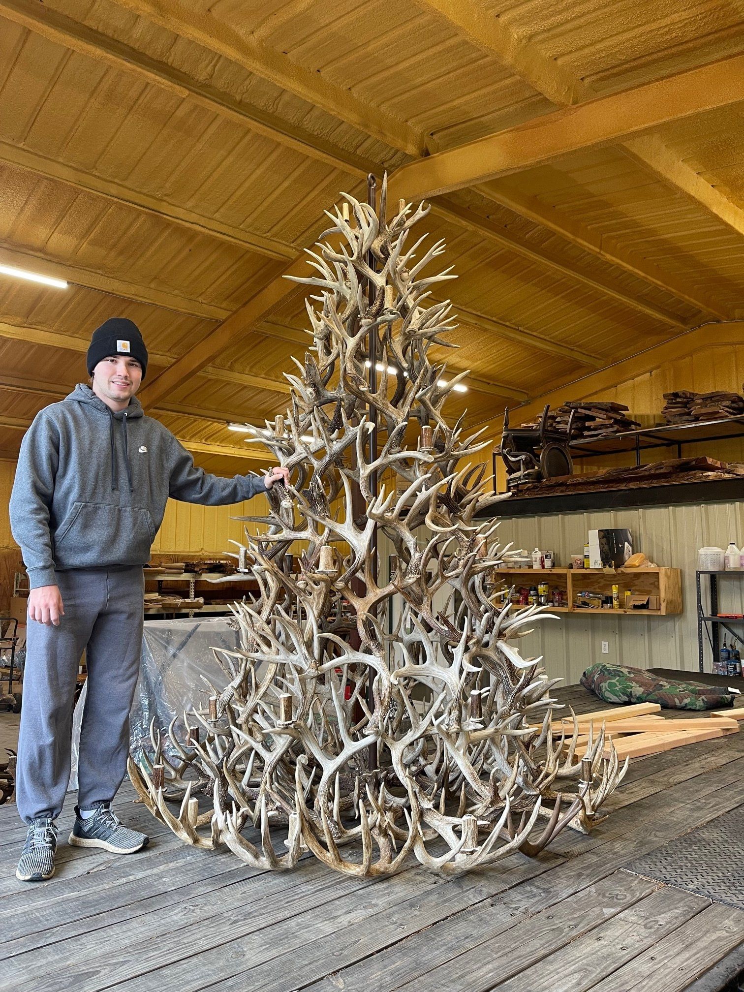 A man is standing next to a large christmas tree made out of deer antlers.