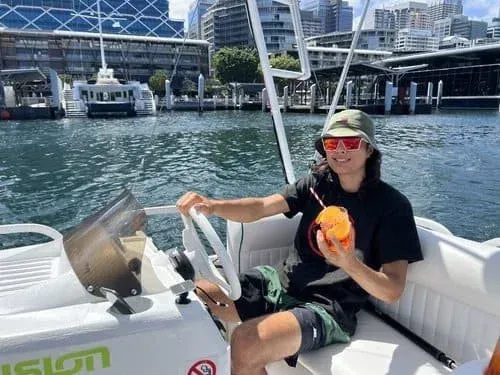 A man is sitting on the steering wheel of a boat holding an orange.
