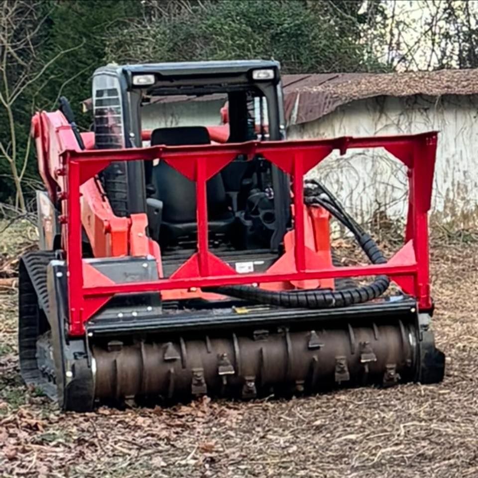 A red skid steer with a forestry mulcher attachment parked on cleared ground near a wooded area.