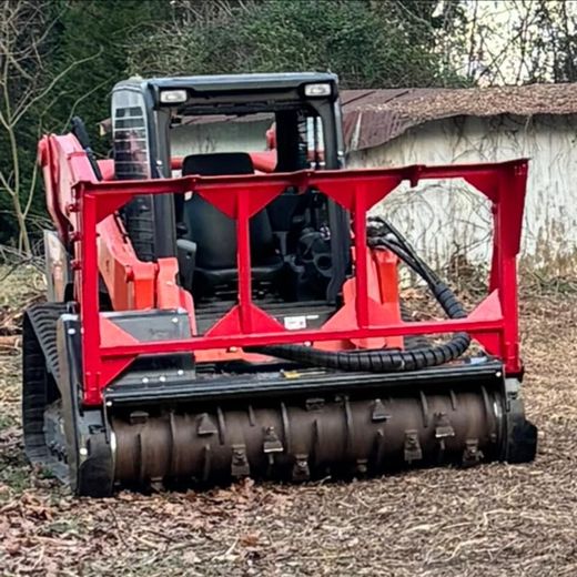 A red skid steer with a forestry mulcher attachment parked on cleared ground near a wooded area.
