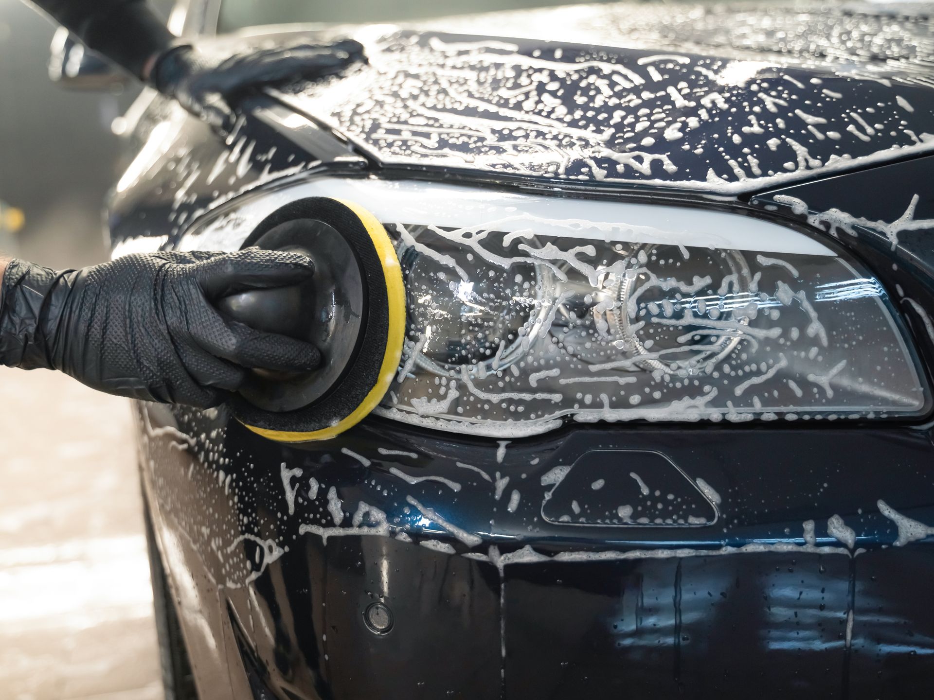 Person polishing a black car headlight with a foam pad and soap suds