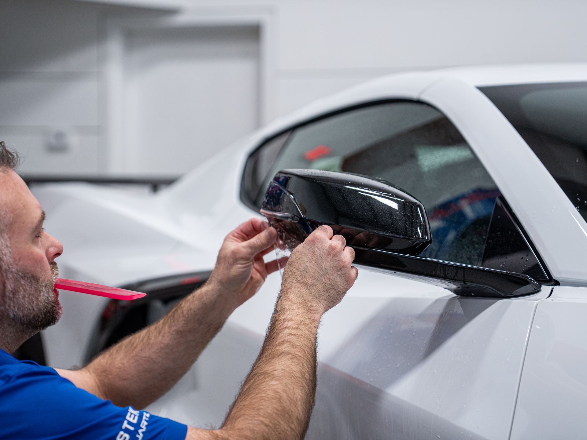 Mechanic applying film to a white car’s side mirror in a workshop