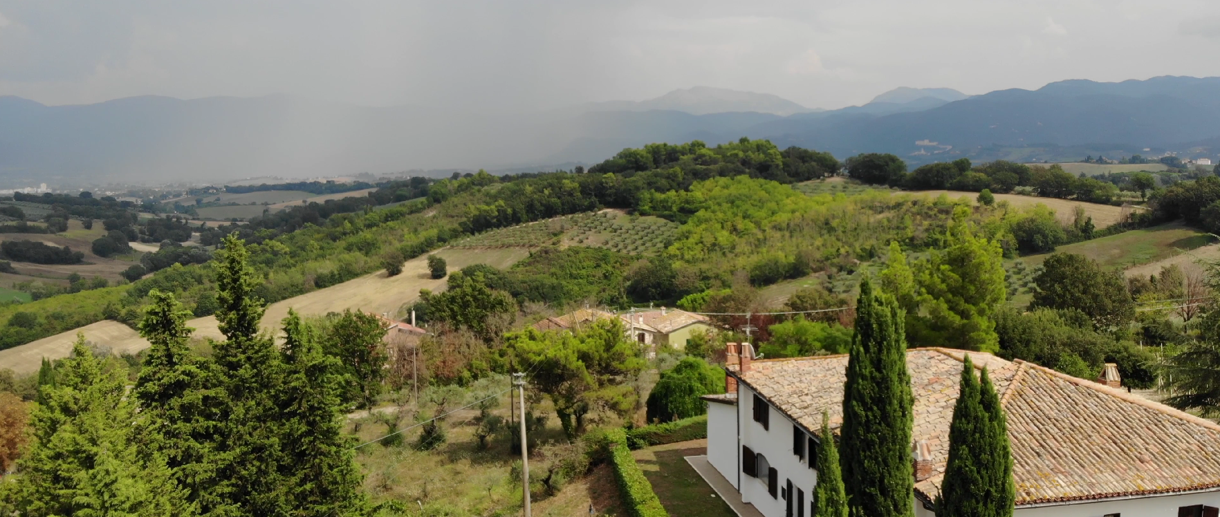 Dolci colline verdi, un edificio bianco con il tetto in terracotta e montagne sotto un cielo coperto.