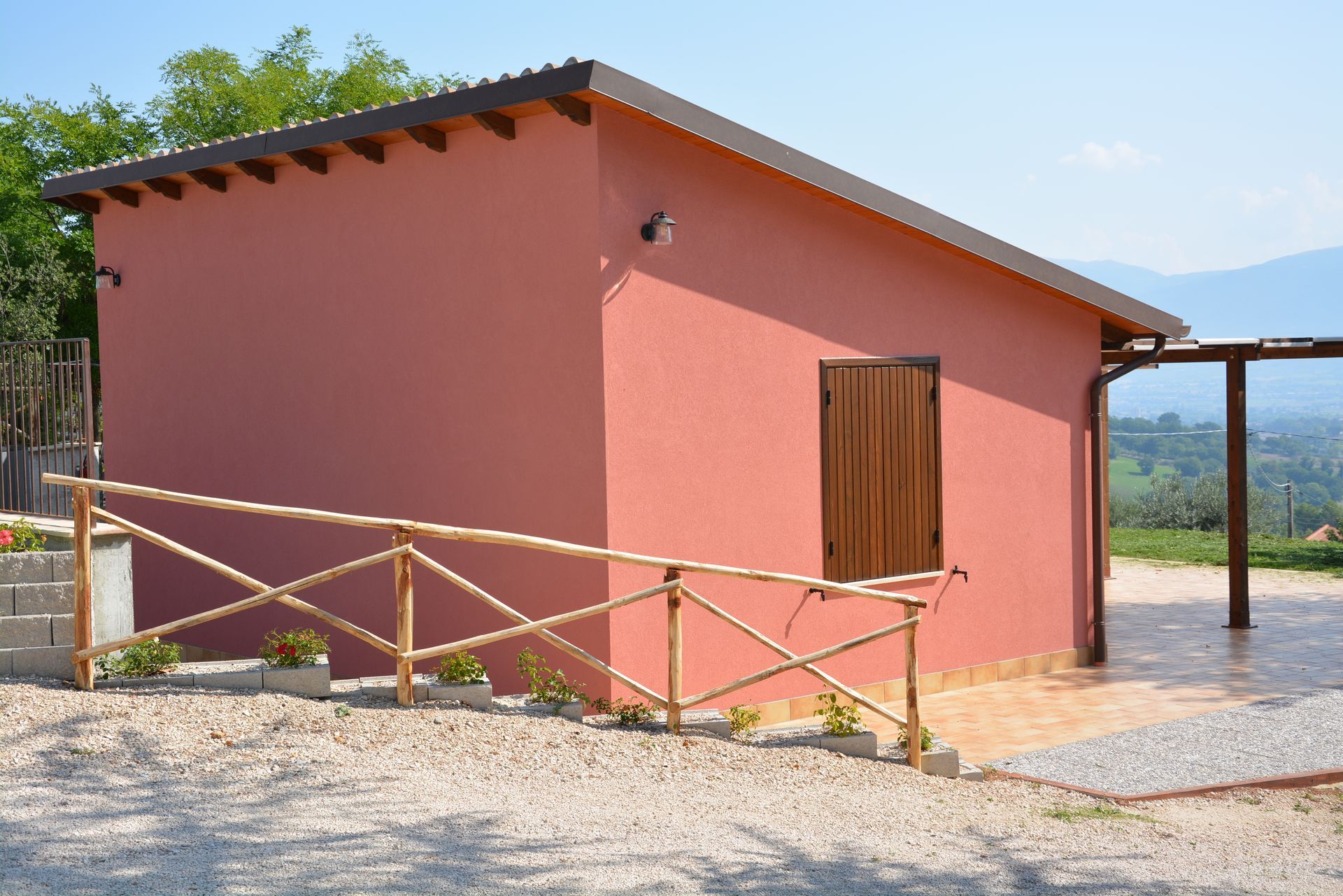 Edificio in stucco rosso con porta in legno, circondato da ghiaia, ringhiera e vista sulle montagne in lontananza.