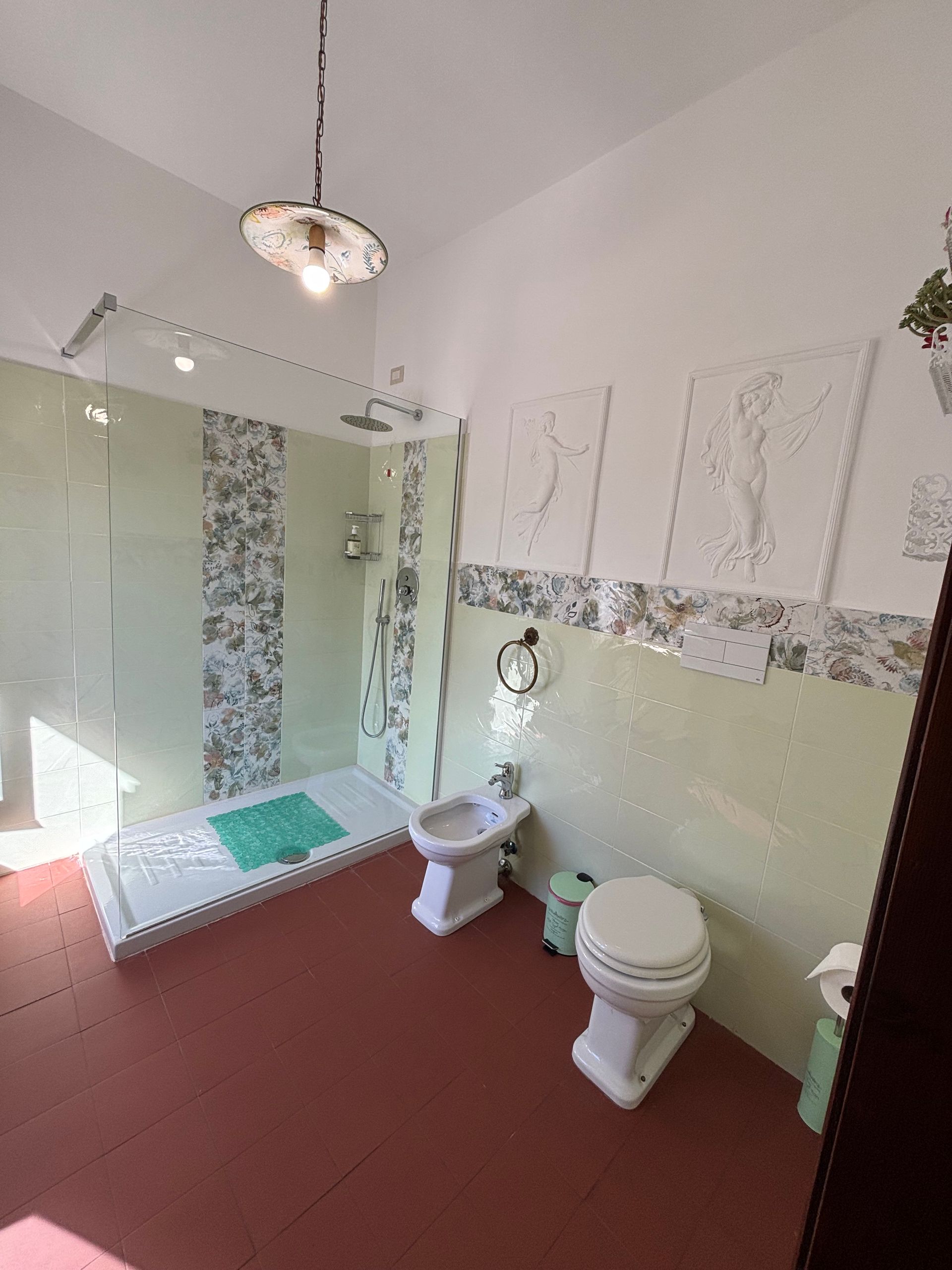 Bagno con doccia, WC e bidet. Pavimento rosso, dettagli in piastrelle verdi e bianche, tre opere d'arte alle pareti.