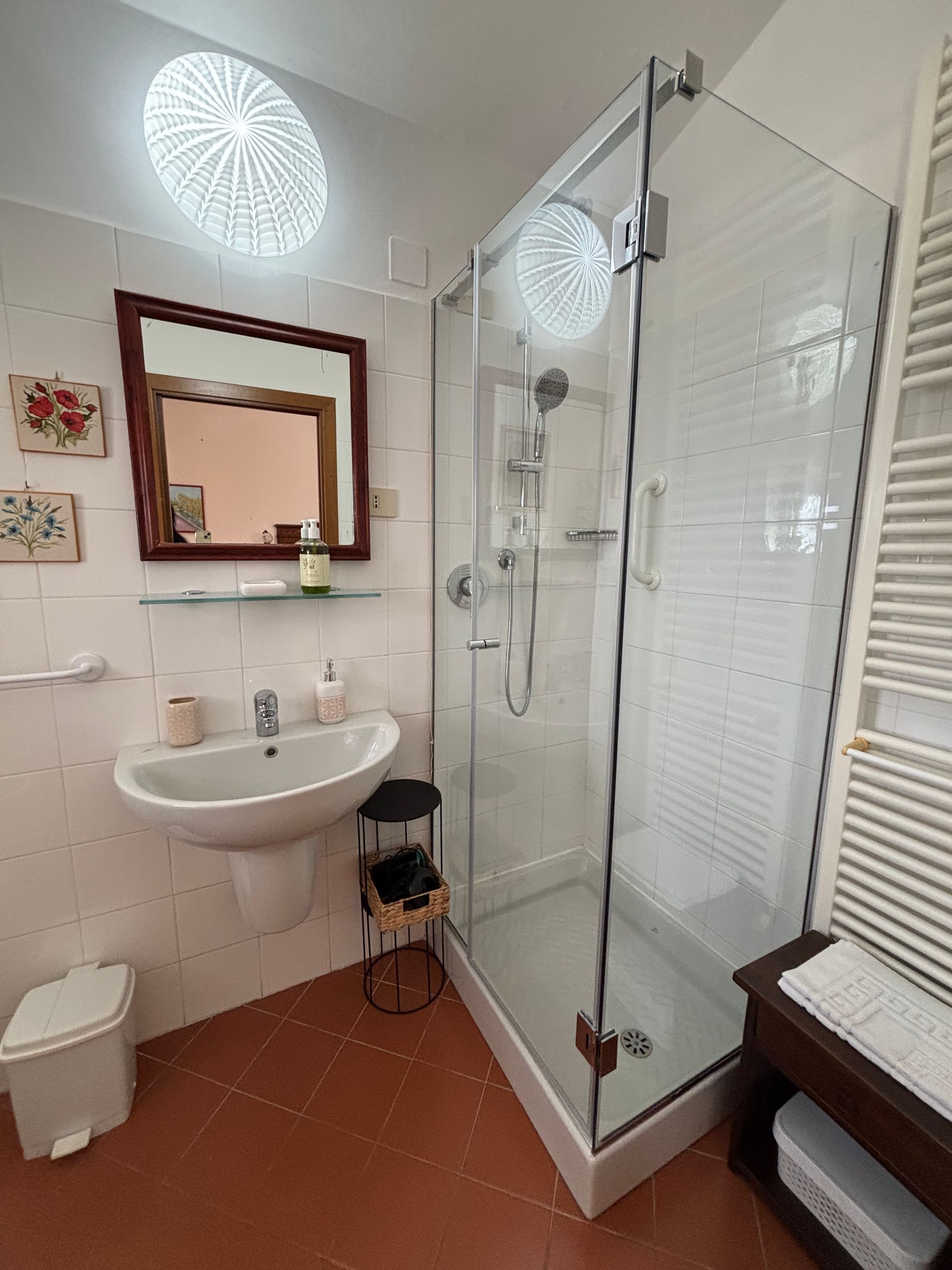 Bagno con doccia in vetro, lavandino, specchio e termosifone. Pareti bianche e pavimento arancione.