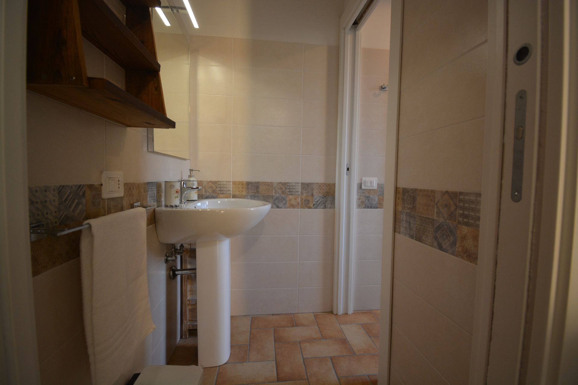 Bagno con lavabo su piedistallo, piastrelle bianche, dettagli marroni e beige, porta aperta sulla doccia.