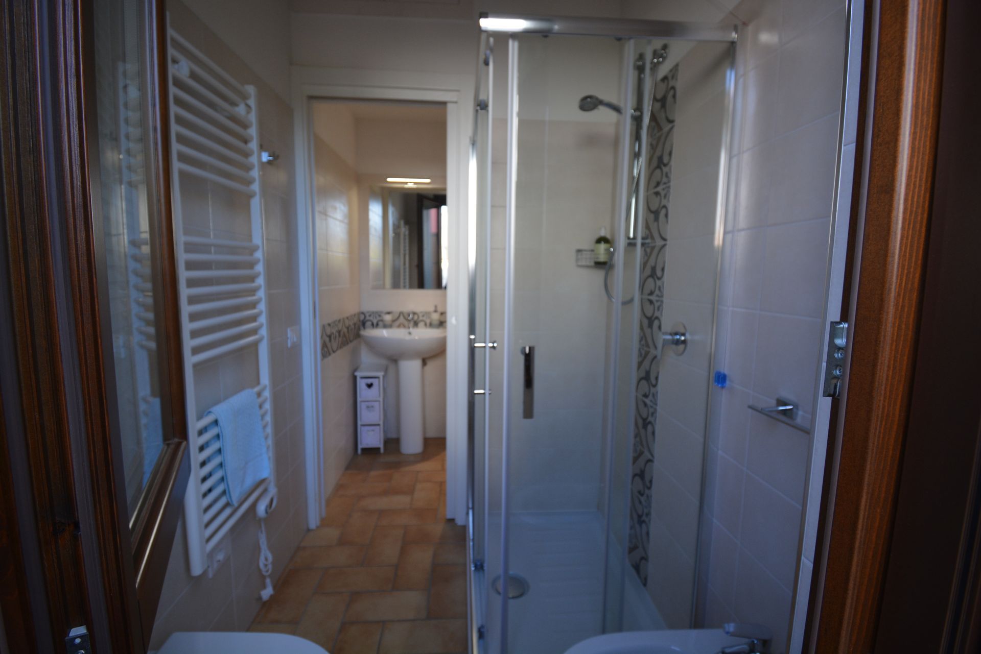 Piccolo bagno con doccia, WC e lavandino a vista. Piastrelle bianche e colori neutri in tutto l'ambiente.