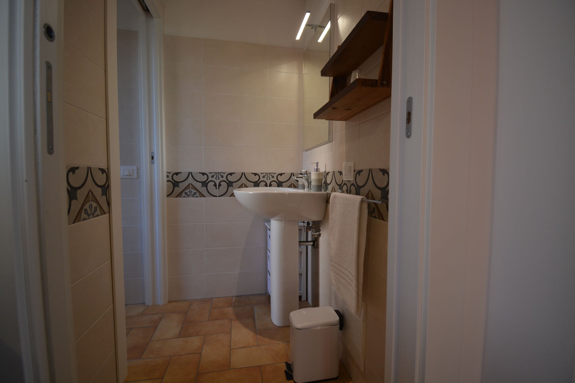 Bagno con lavabo su piedistallo, paraschizzi decorativo in piastrelle, asciugamano, mensola in legno e pareti bianche.