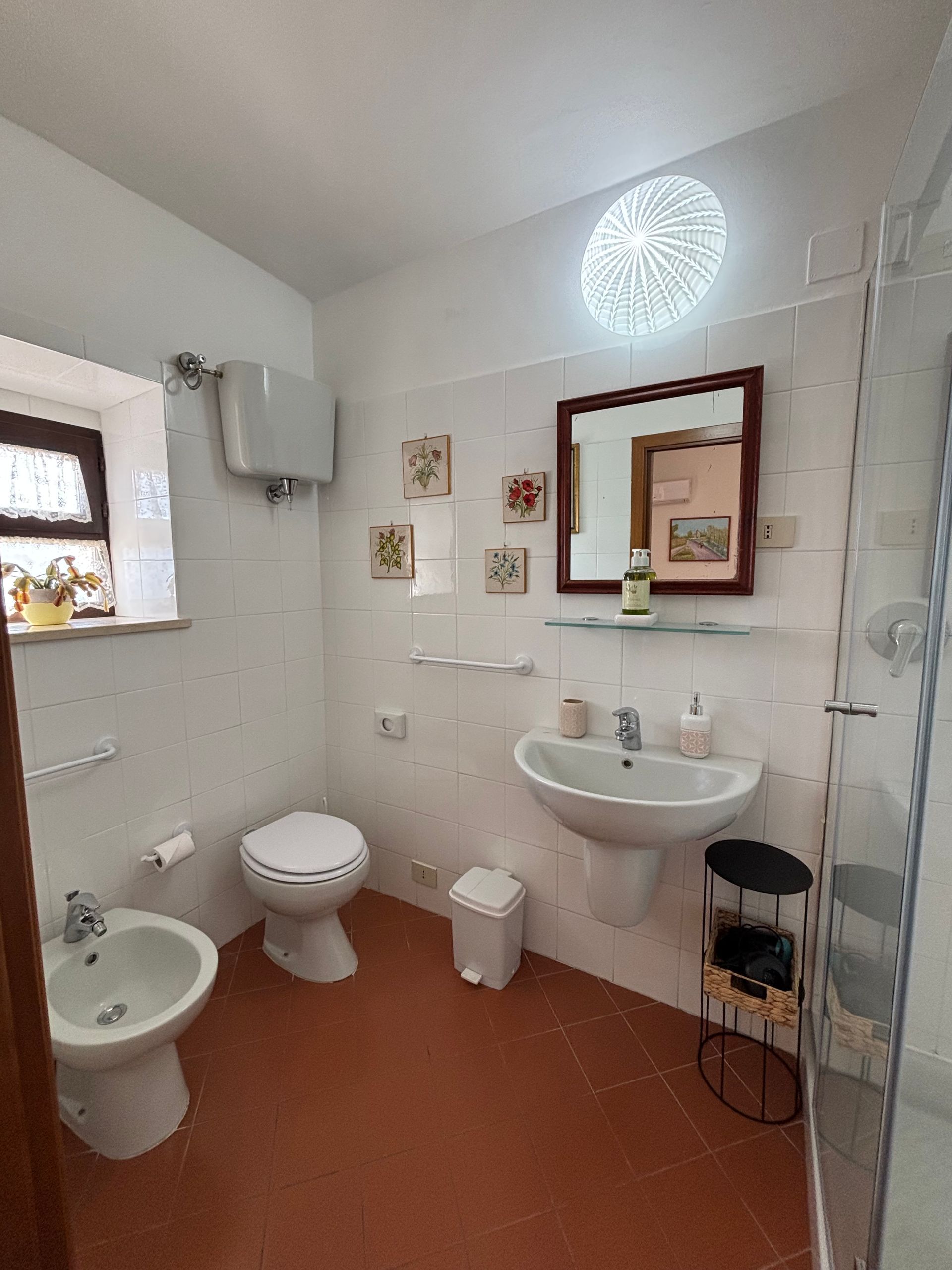 Bagno con WC, bidet, lavandino, doccia e piccola finestra. Piastrelle bianche, pavimento rosso e opere d'arte decorative.