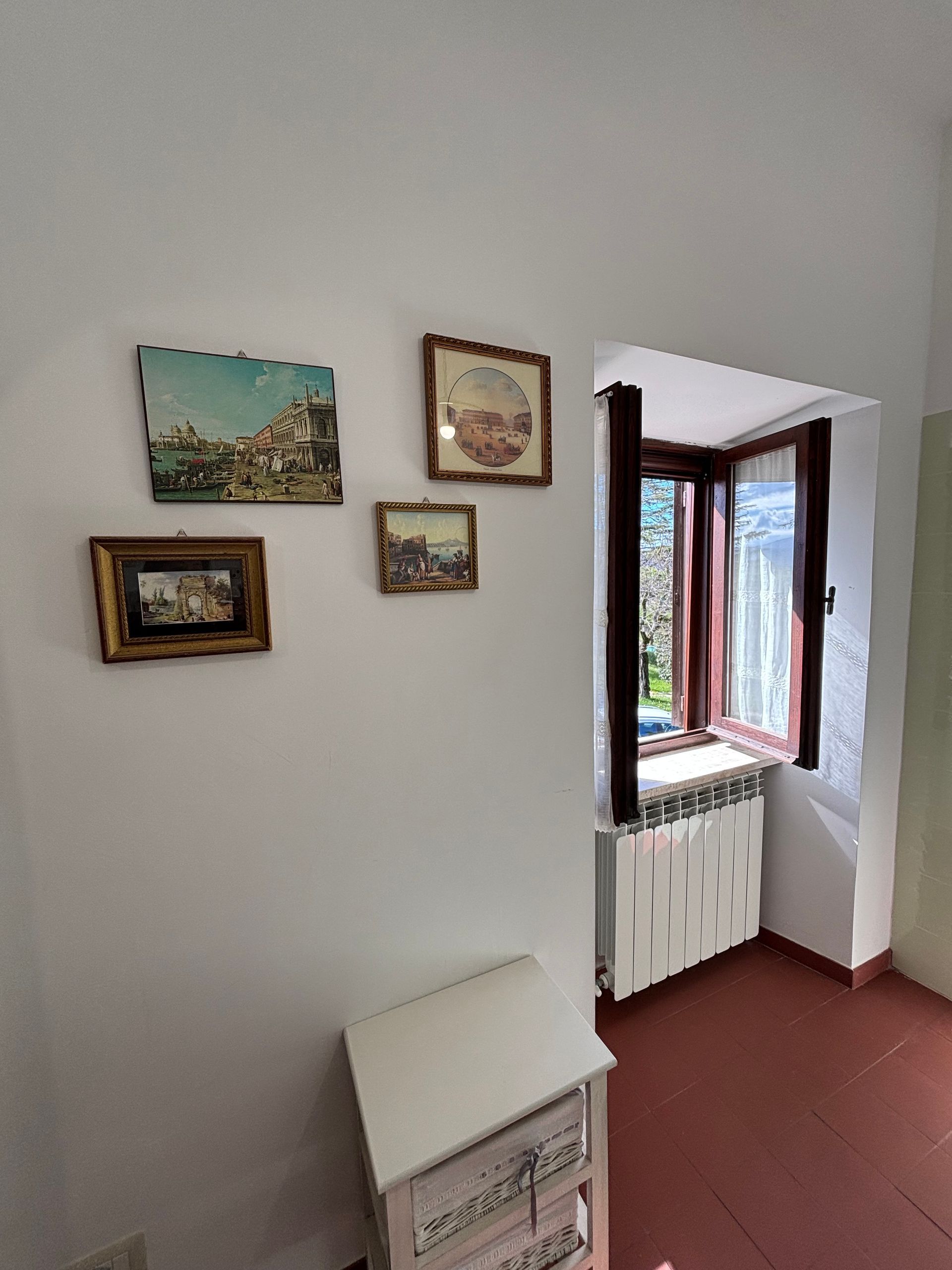 Parete bianca con un quadro incorniciato sopra una piccola cassapanca; una finestra con cornice scura e un termosifone sotto.