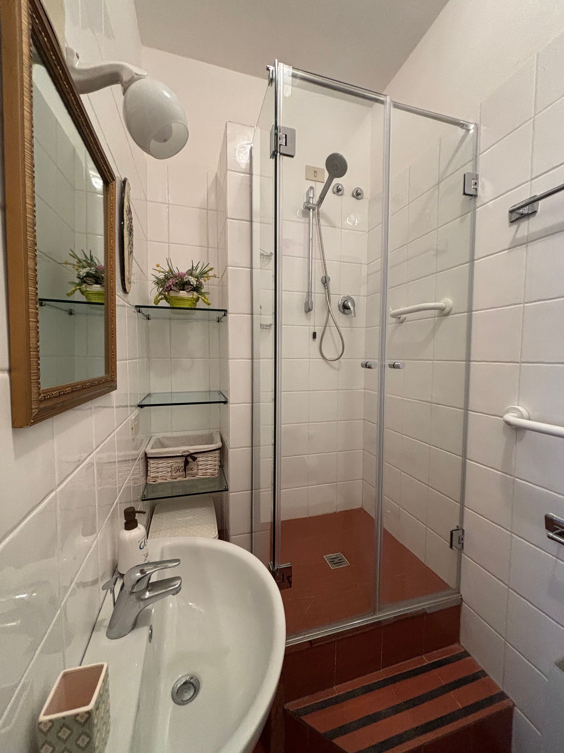 Piccolo bagno con doccia angolare, lavandino e specchio. Pareti piastrellate bianche con ripiani in vetro.