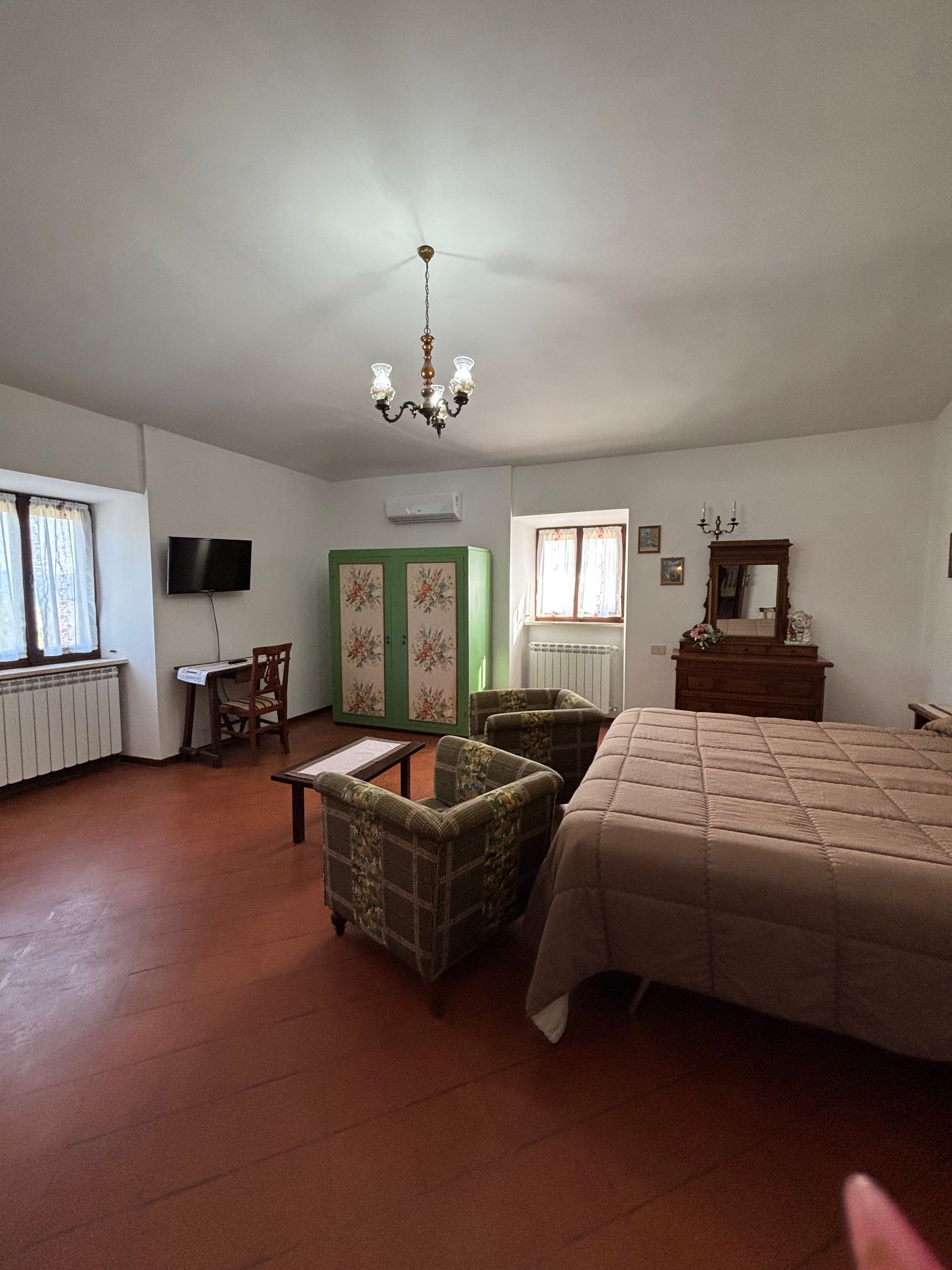 Ampia camera da letto con letto, due poltrone, un mobile dipinto, TV e lampadario. Pavimento rosso, pareti bianche.