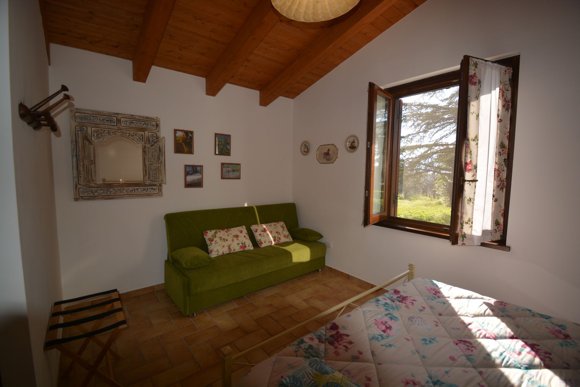 Accogliente camera da letto con divano verde, copriletto floreale e finestra con vista su un paesaggio verde.
