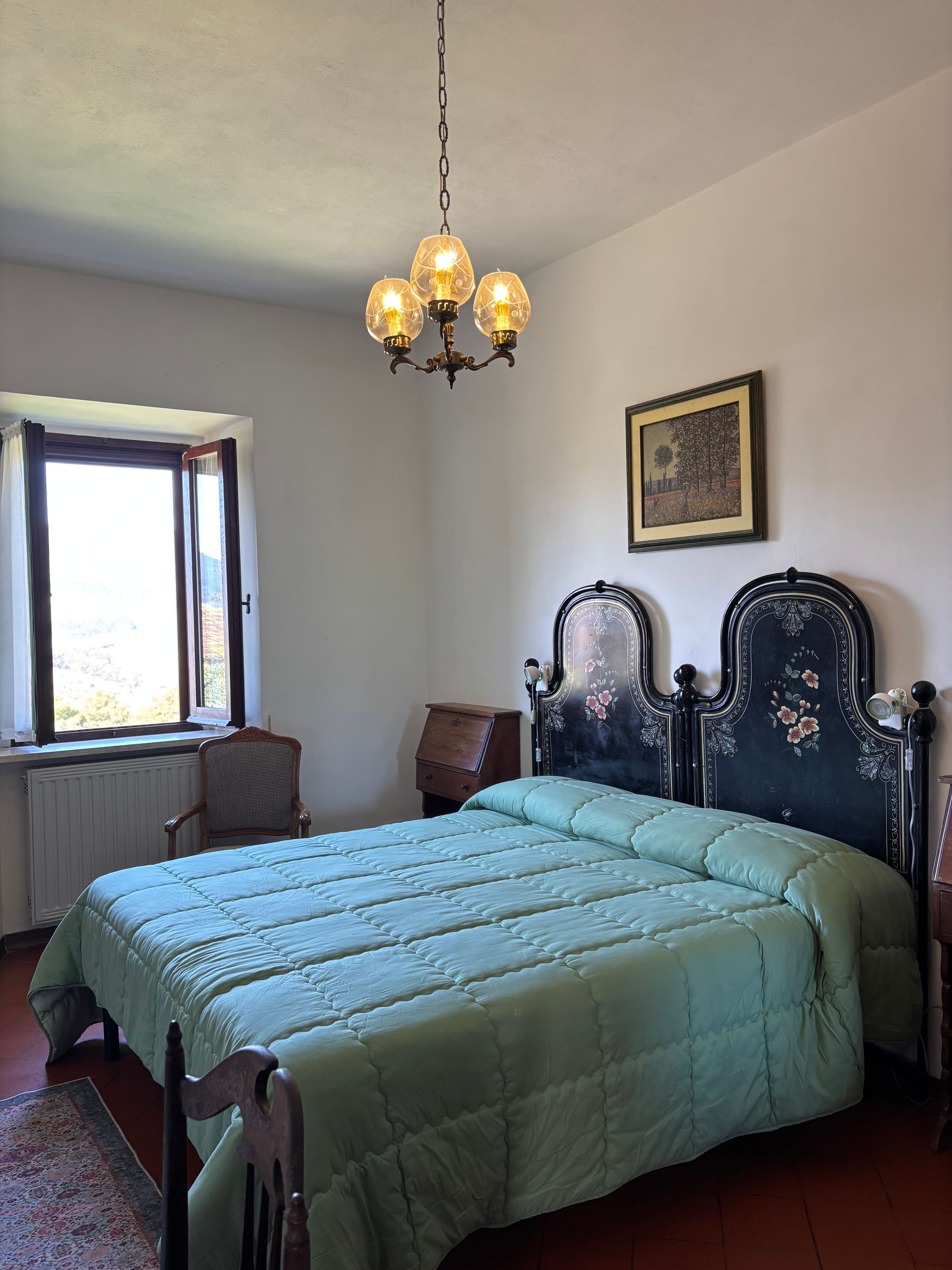 Camera da letto con letto matrimoniale, testiera antica, finestra, lampadario e piccole opere d'arte incorniciate.