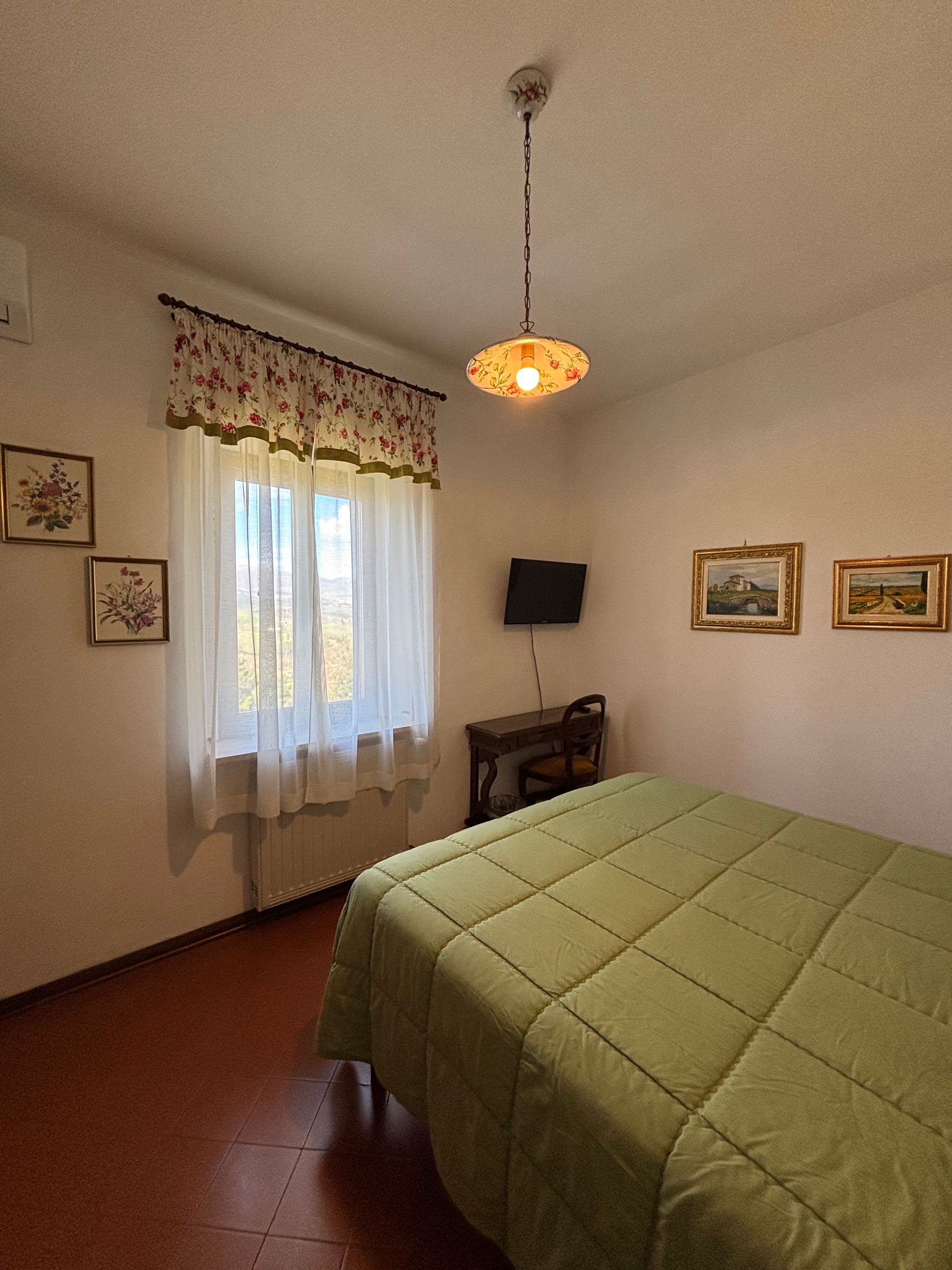 Camera da letto con copriletto verde, piccola TV, finestra con balza floreale e quadri alle pareti.