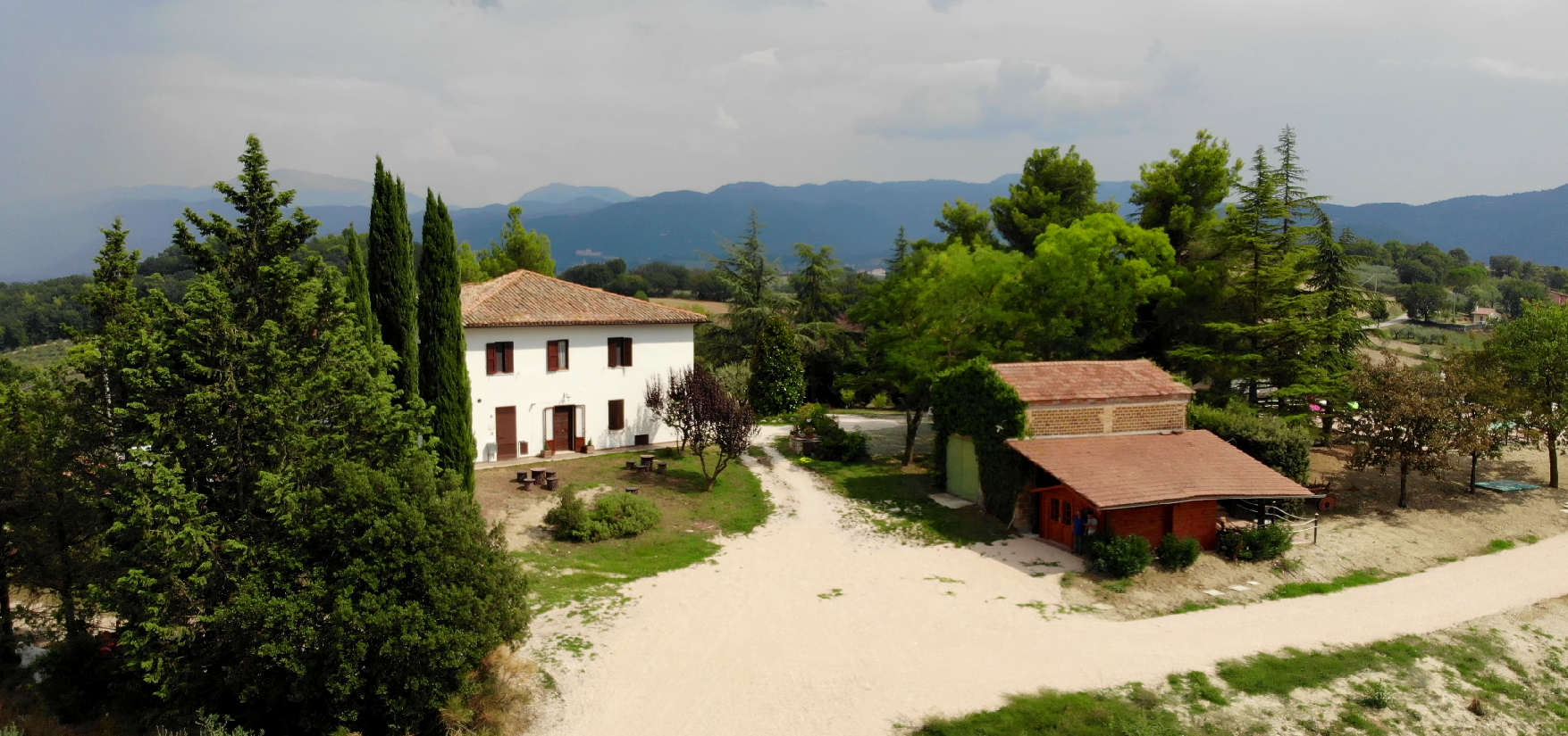 Una vista aerea di una casa in stucco bianco e di un edificio con il tetto rosso, circondati da alberi, in una giornata di sole.