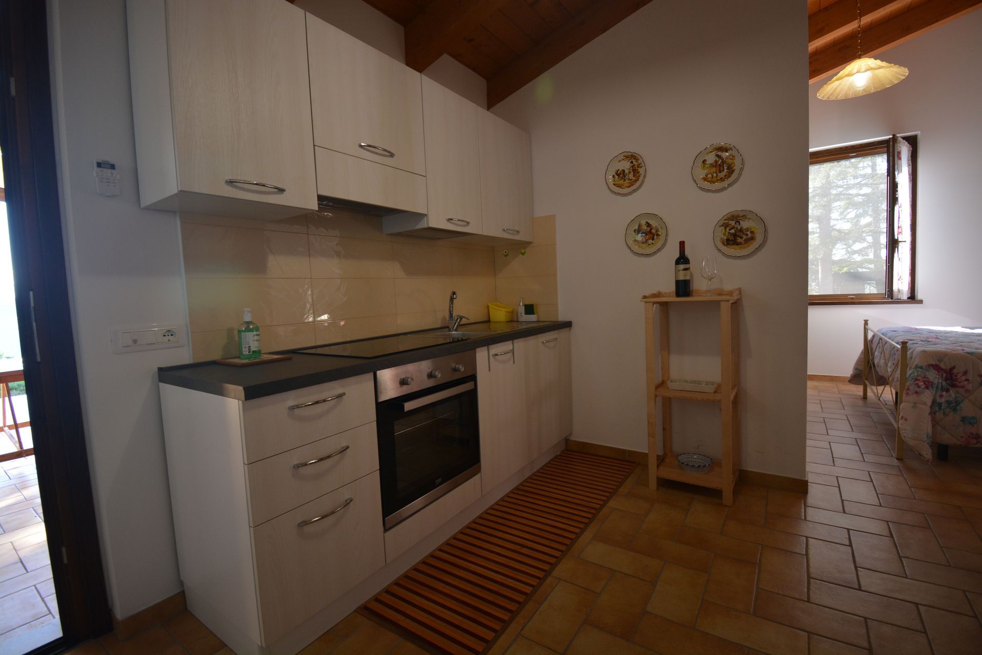 Piccola cucina con mobili bianchi, forno e mensola in legno; camera da letto adiacente con letto.