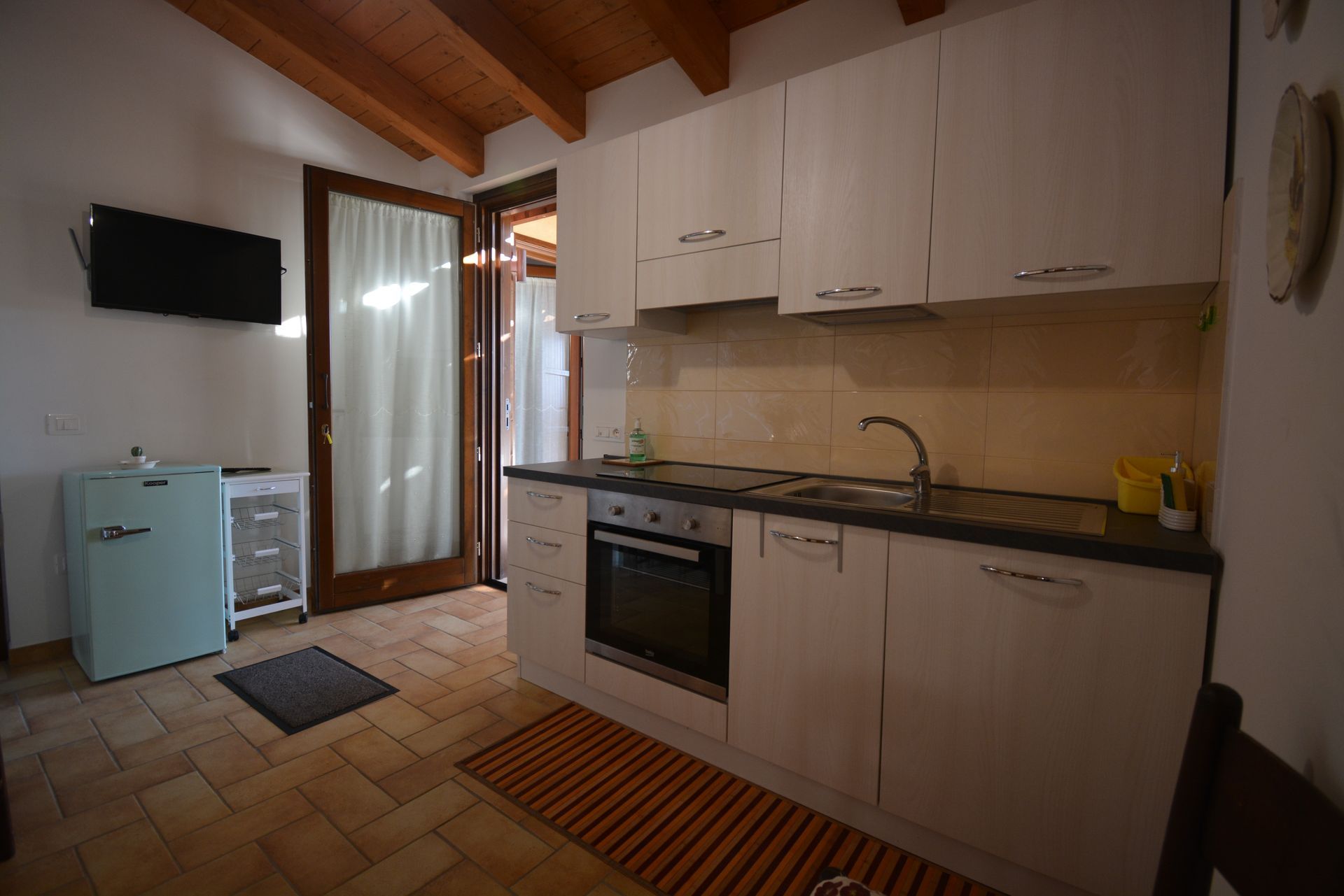 Cucina con mobili bianchi, soffitto in legno chiaro, frigorifero, forno e porta d'accesso all'esterno.