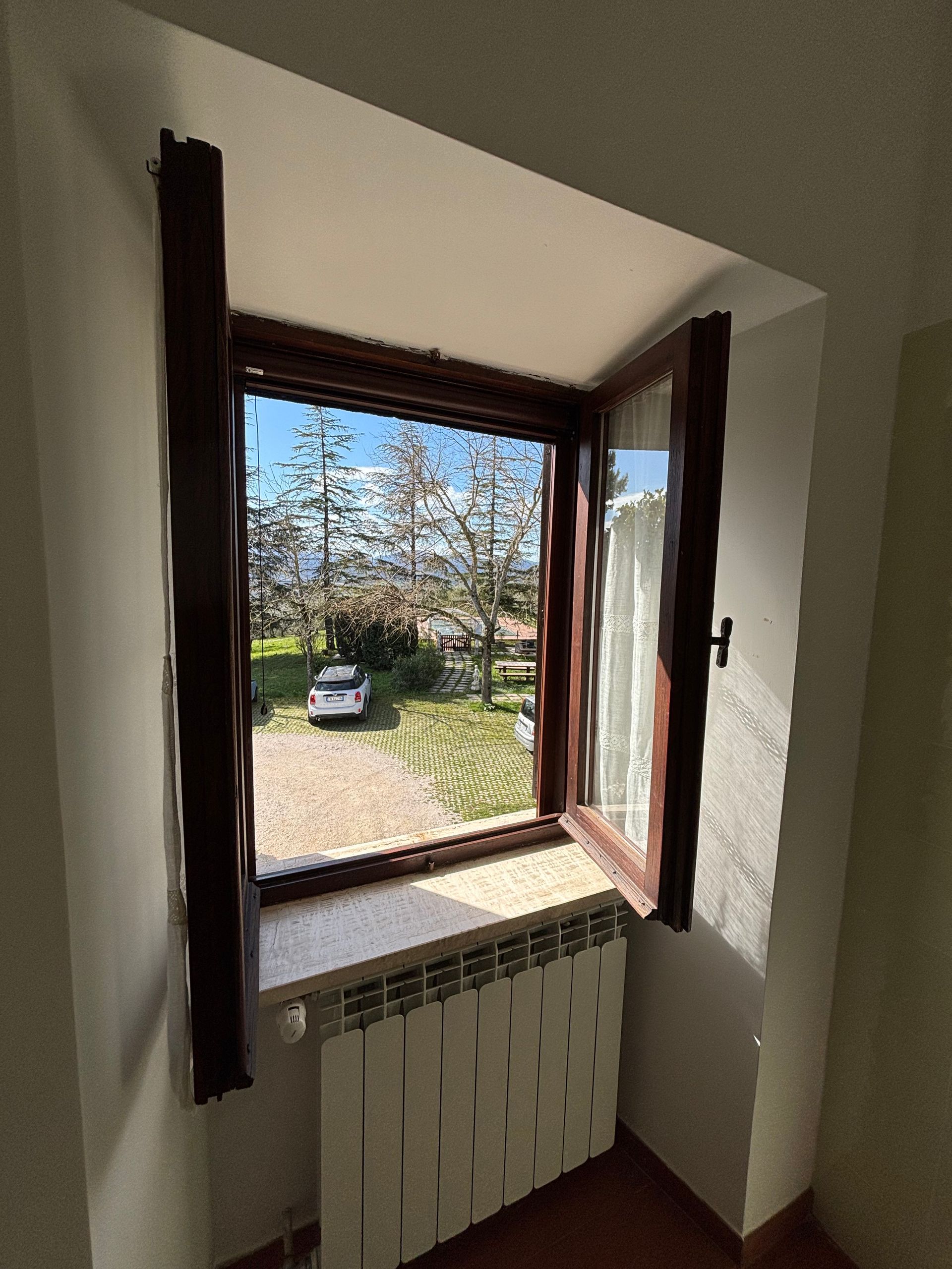Finestra in legno aperta con vista sul cortile e sull'auto, radiatore bianco sotto.