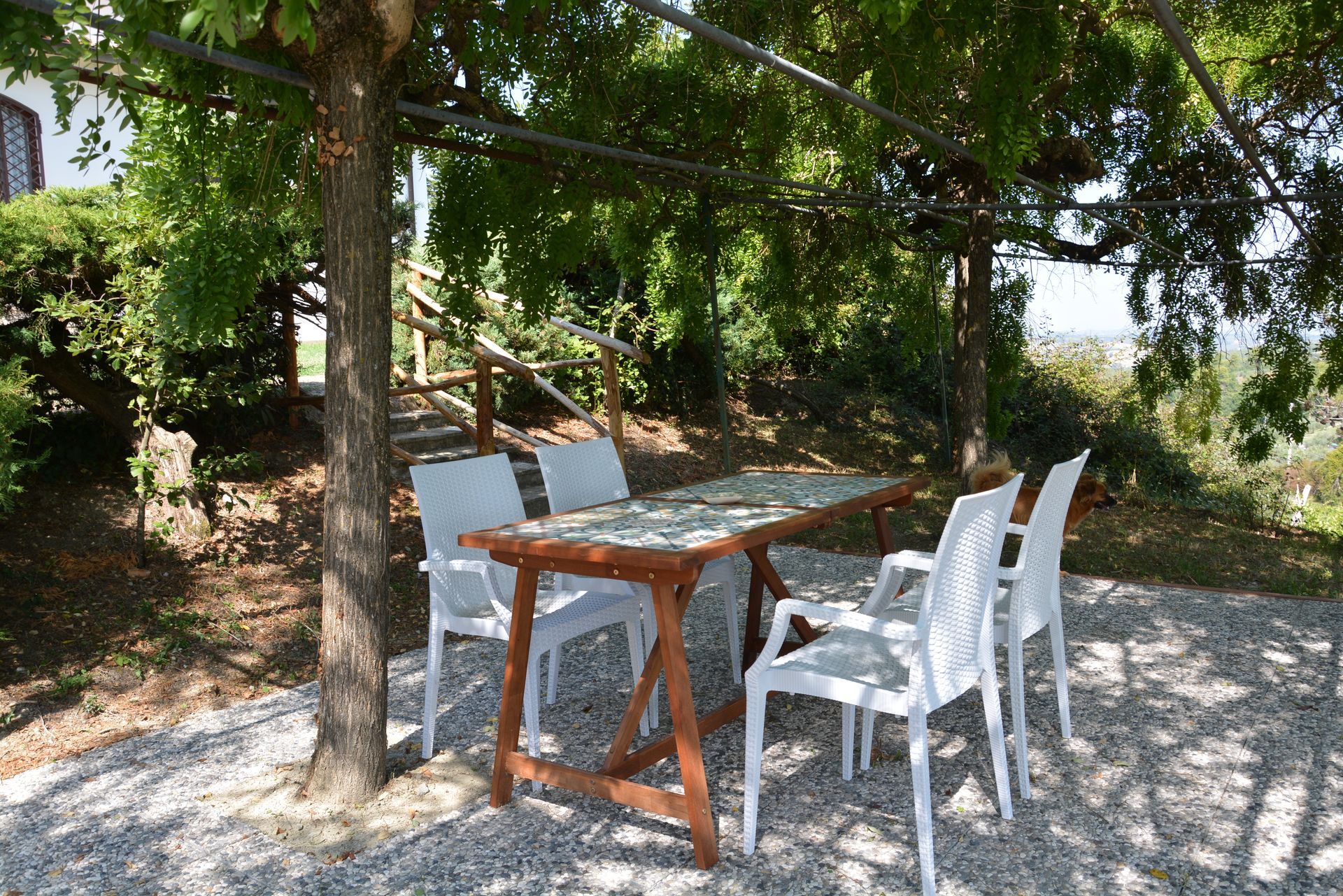 Tavolo e sedie da esterno sotto un pergolato ombreggiato in una zona ghiaiosa.