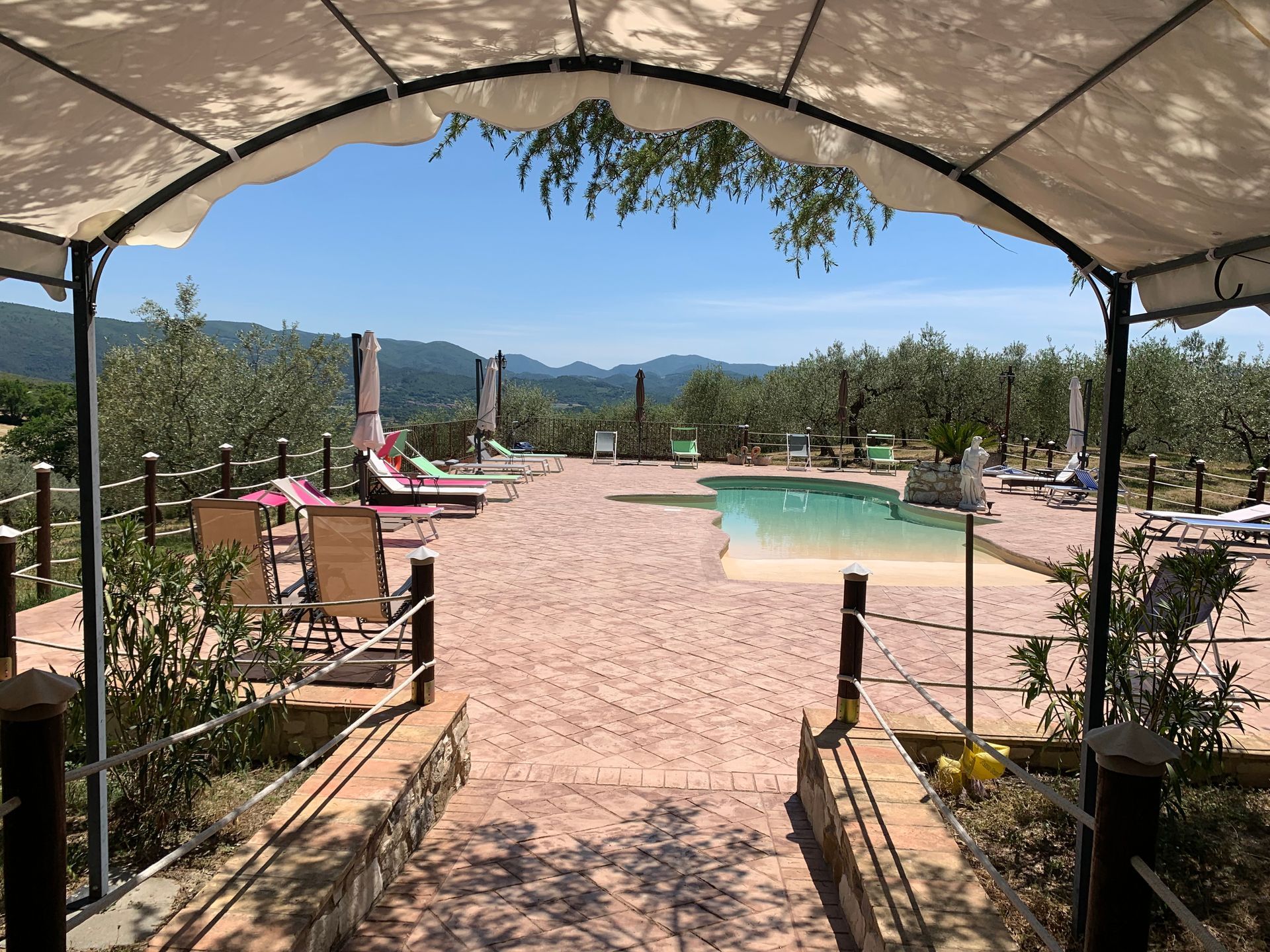 Area piscina con sedie a sdraio, alberi e vista sulle montagne, incorniciata da una tettoia ad arco.