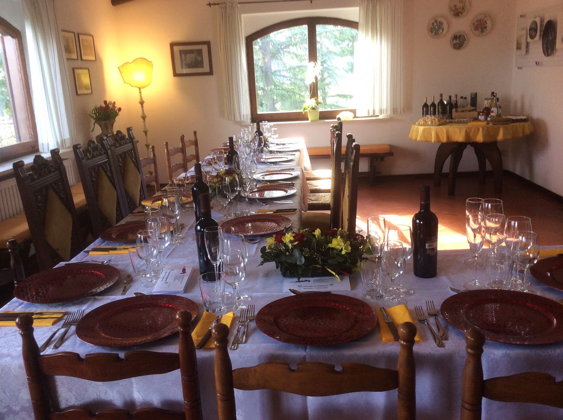 Tavolo da pranzo apparecchiato per un pasto, con piatti marroni, bicchieri da vino e un centrotavola floreale in una stanza illuminata dal sole.