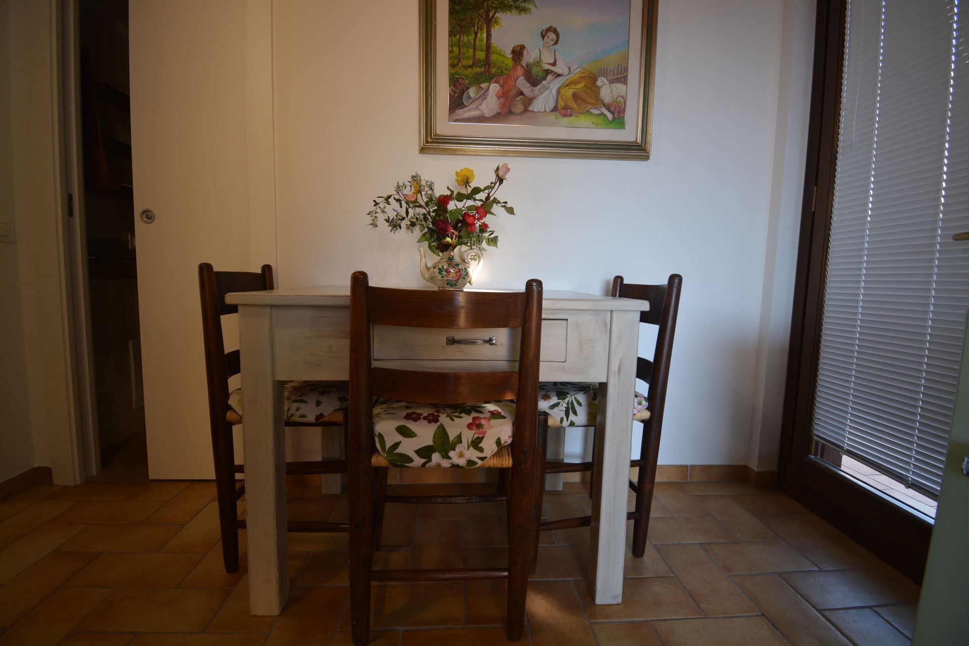 Tavolo da pranzo con tre sedie, fiori e un dipinto su una parete bianca.