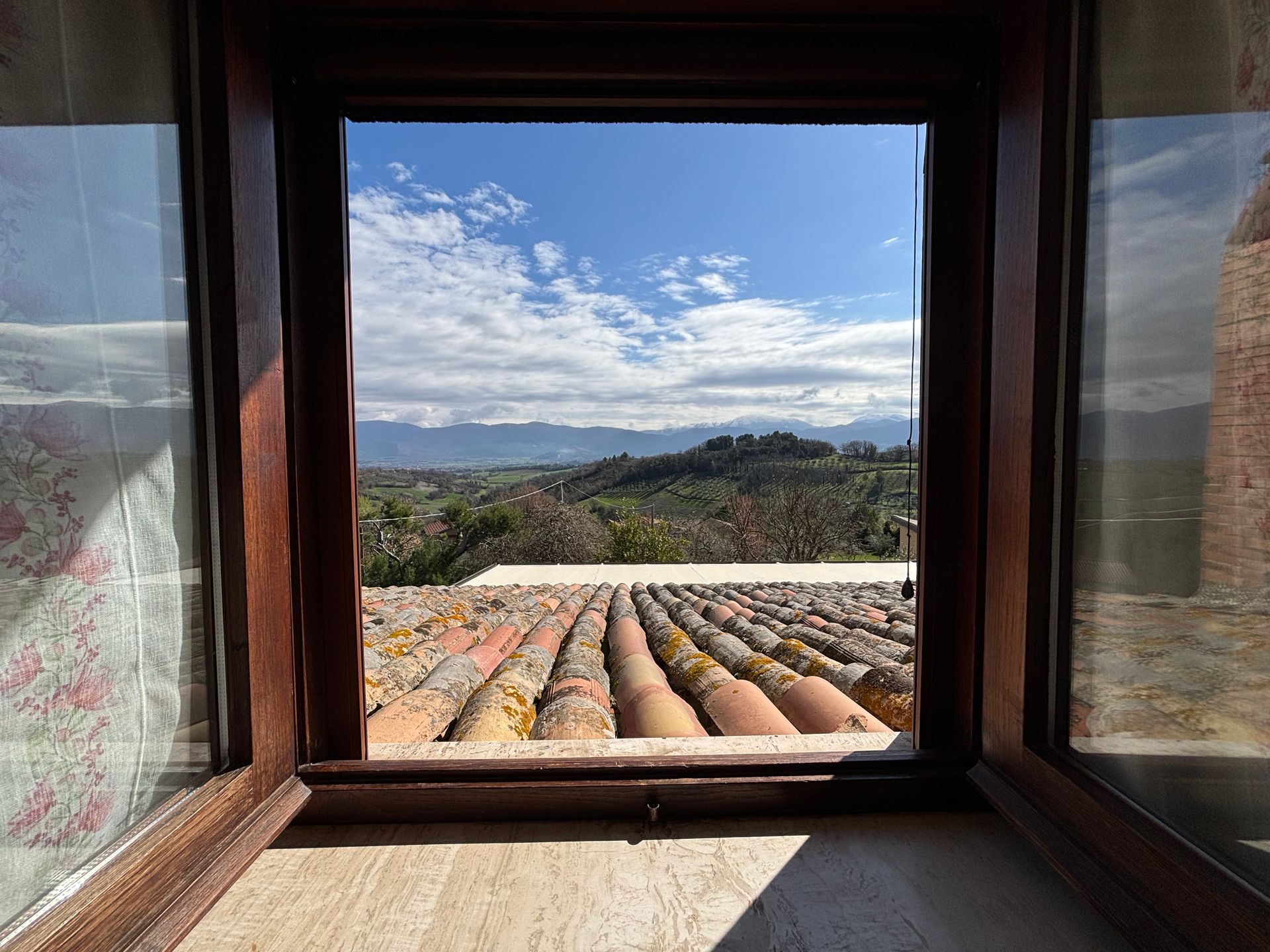 Vista dalla finestra: cielo azzurro, nuvole, montagne, tetto di tegole, struttura in legno, luce del sole.