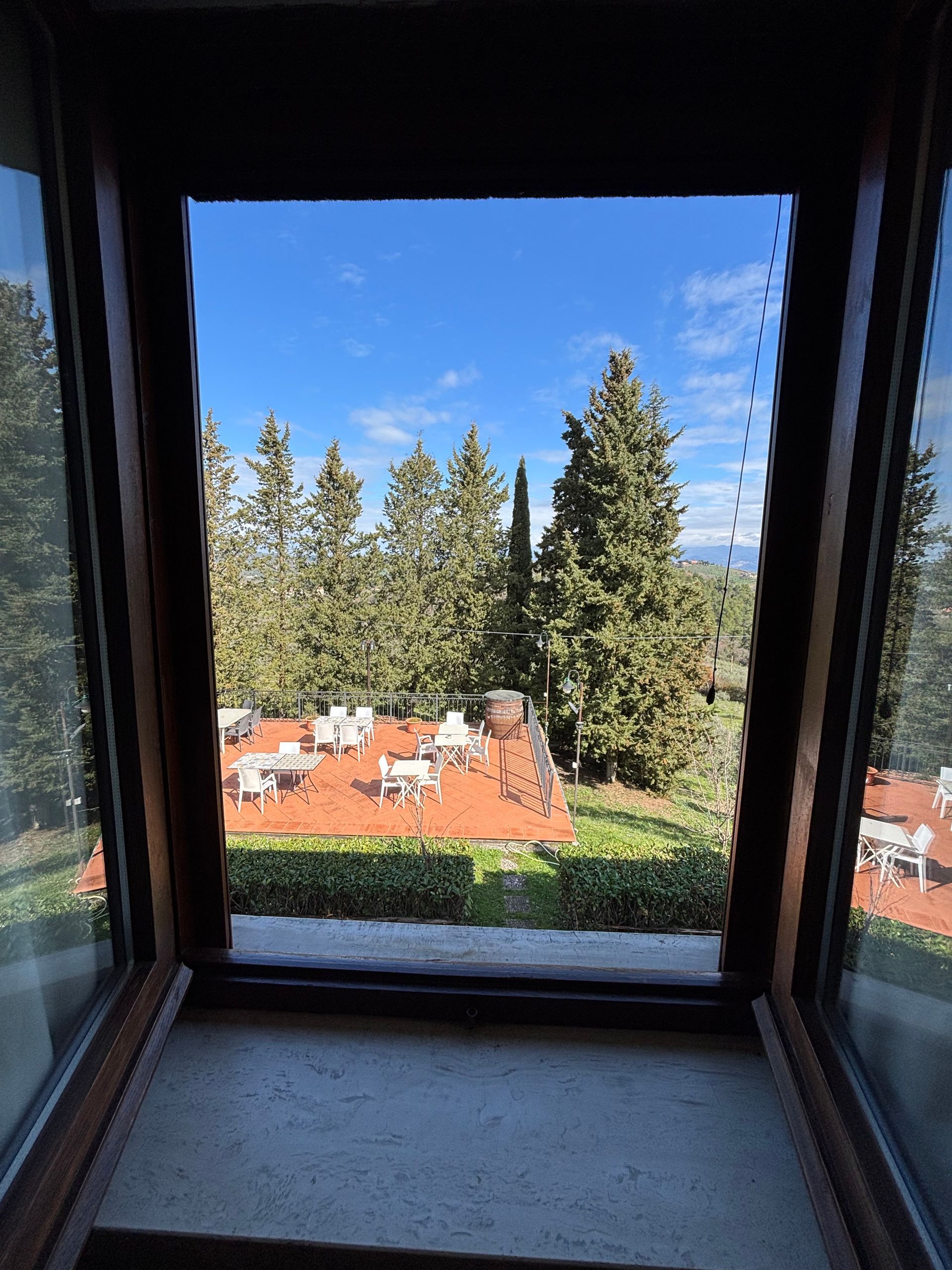 Vista dalla finestra: patio piastrellato di rosso con tavoli e sedie bianchi, alberi, cielo azzurro.