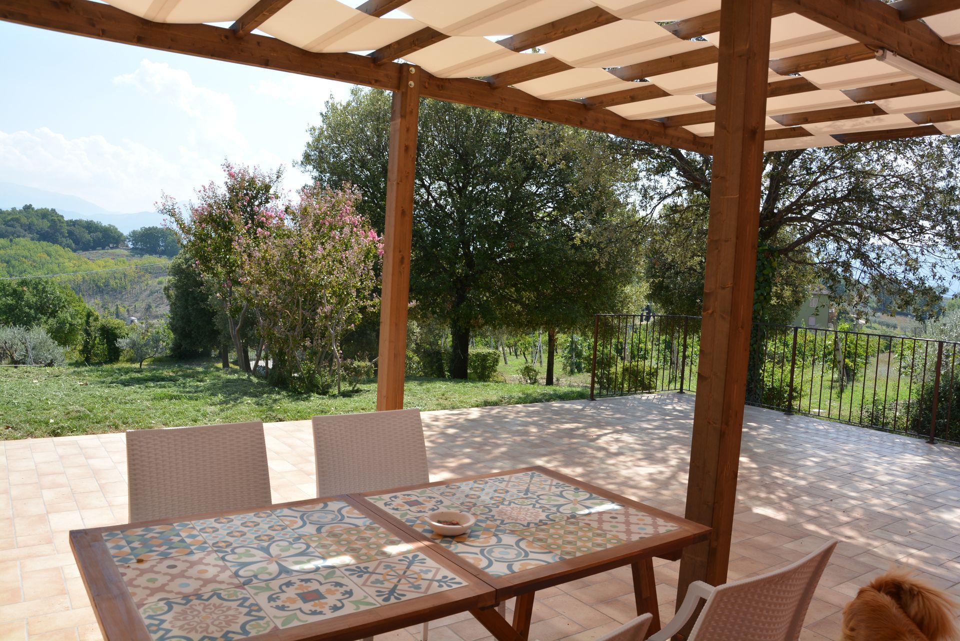 Patio con tavolo piastrellato e sedie sotto un pergolato in legno, con vista sul paesaggio verde.