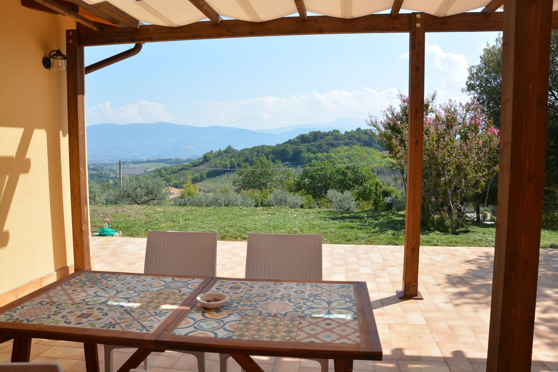 Tavolo da giardino con vista sulle dolci colline verdi sotto un cielo azzurro, incorniciato da un pergolato in legno.