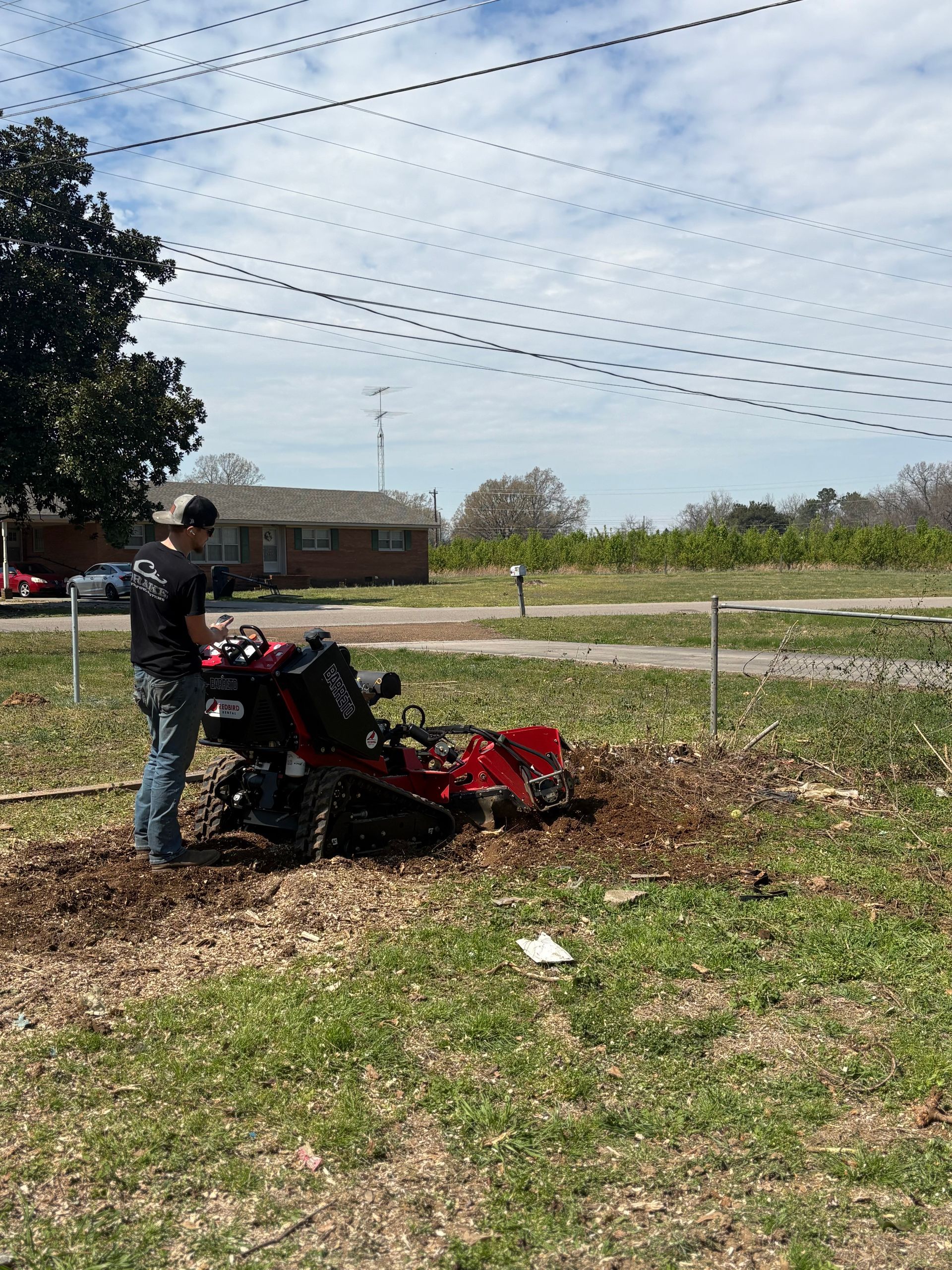 A person operates a red stump grinder in a grassy yard under a bright, cloudy sky with power lines overhead.