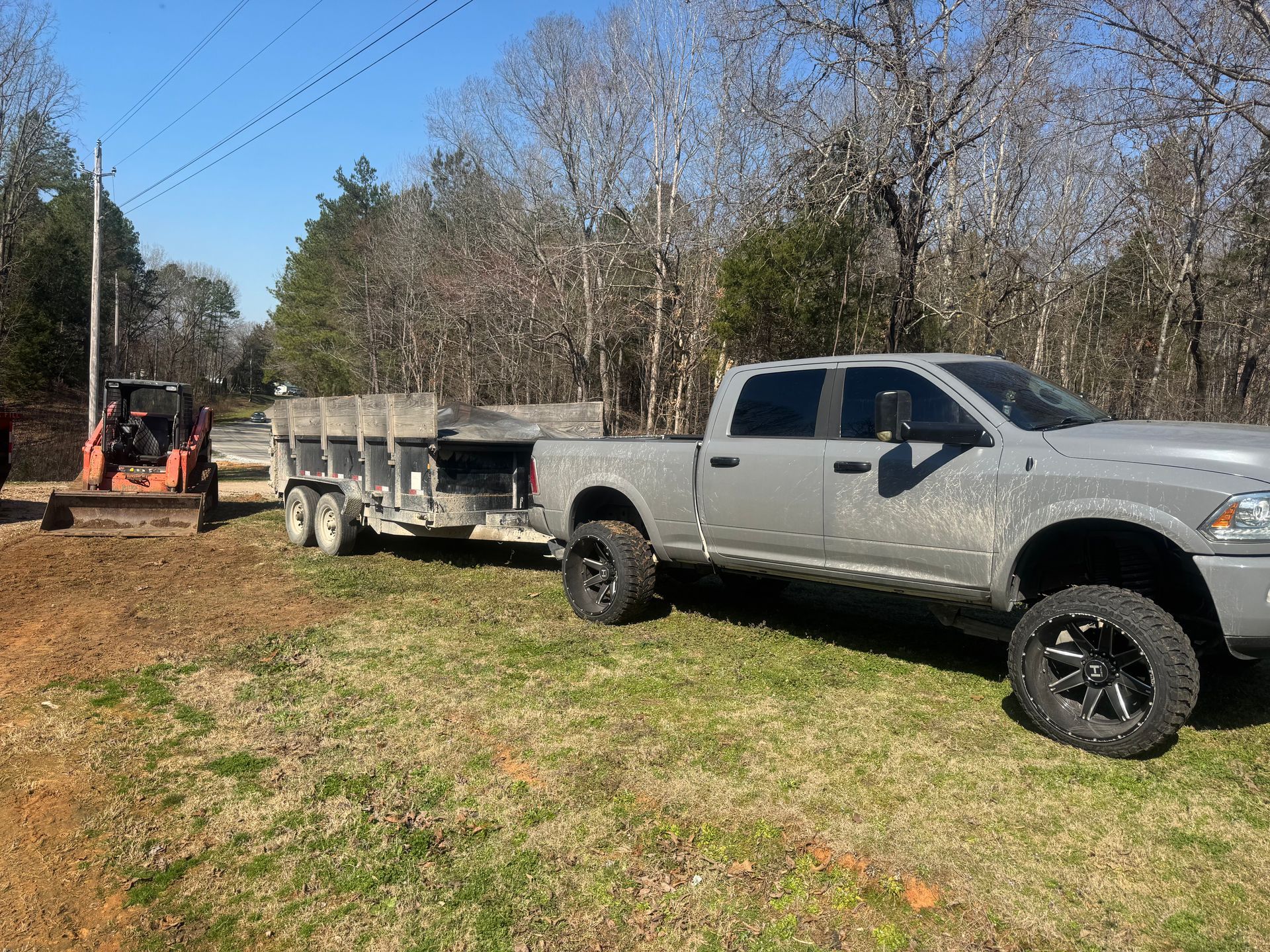 A gray pickup truck towing a flatbed trailer on a grassy lot with an orange skid steer parked nearby.