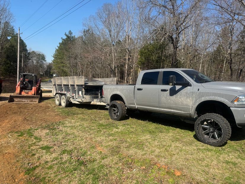 A gray pickup truck towing a flatbed trailer on a grassy lot with an orange skid steer parked nearby.